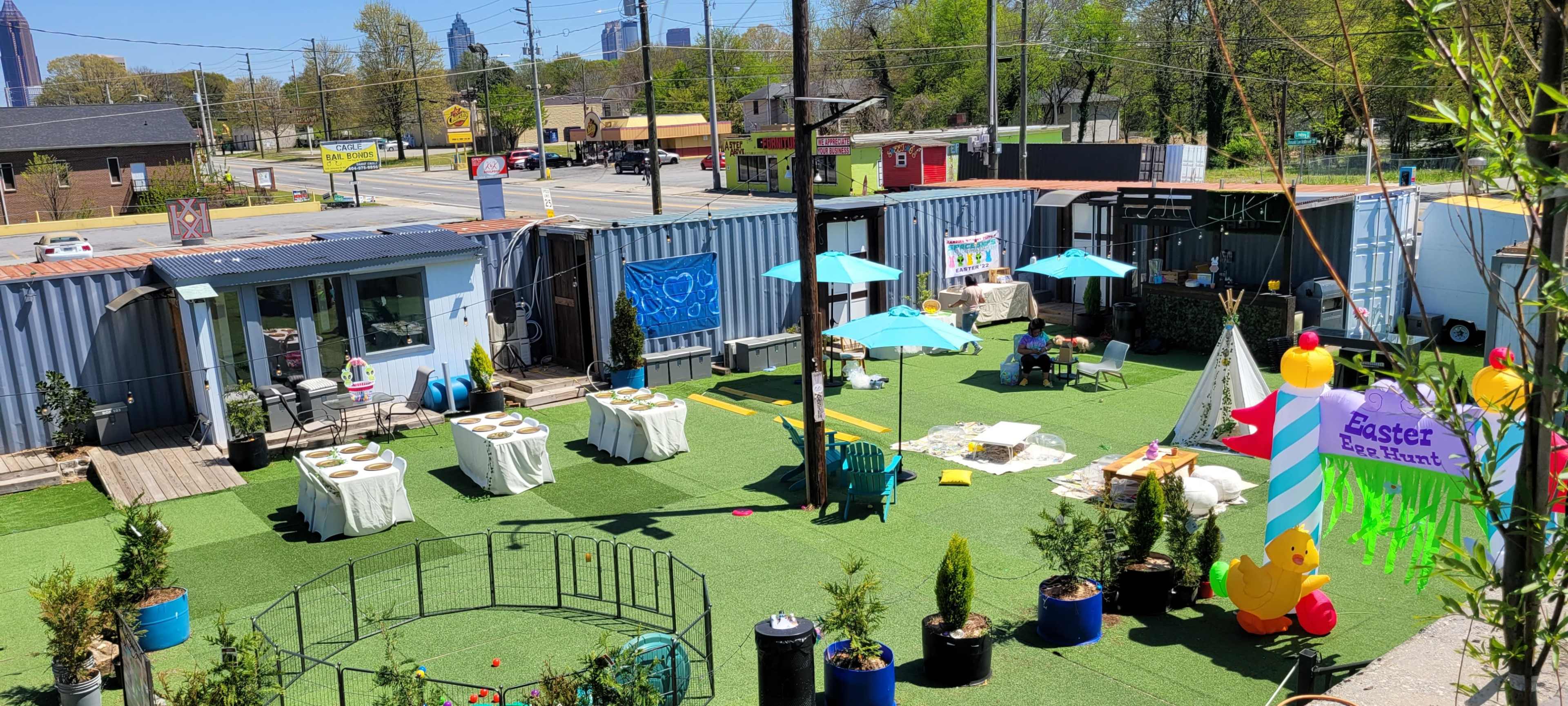An outdoor event space with several tables arranged on green turf, colorful umbrellas, and decorations for an Easter egg hunt.