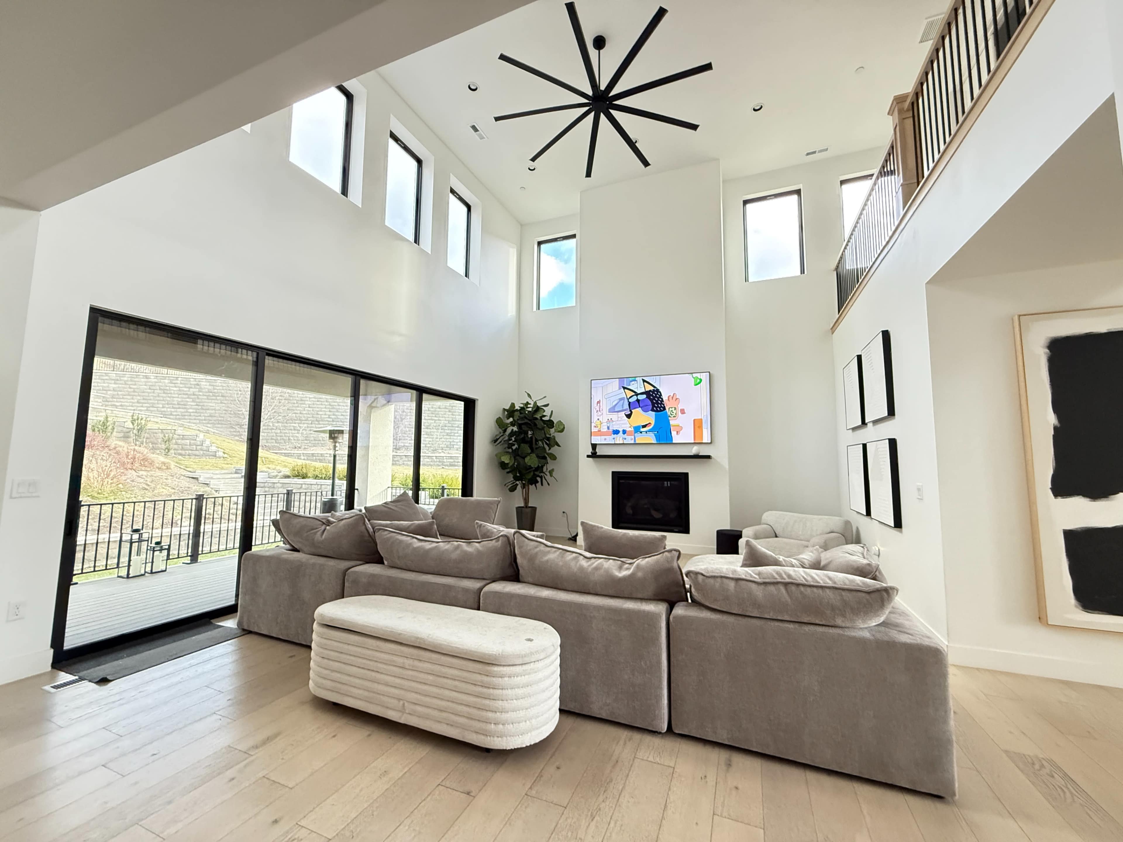 A spacious living room features a large sectional sofa, a wall-mounted television, and floor-to-ceiling windows that provide natural light.