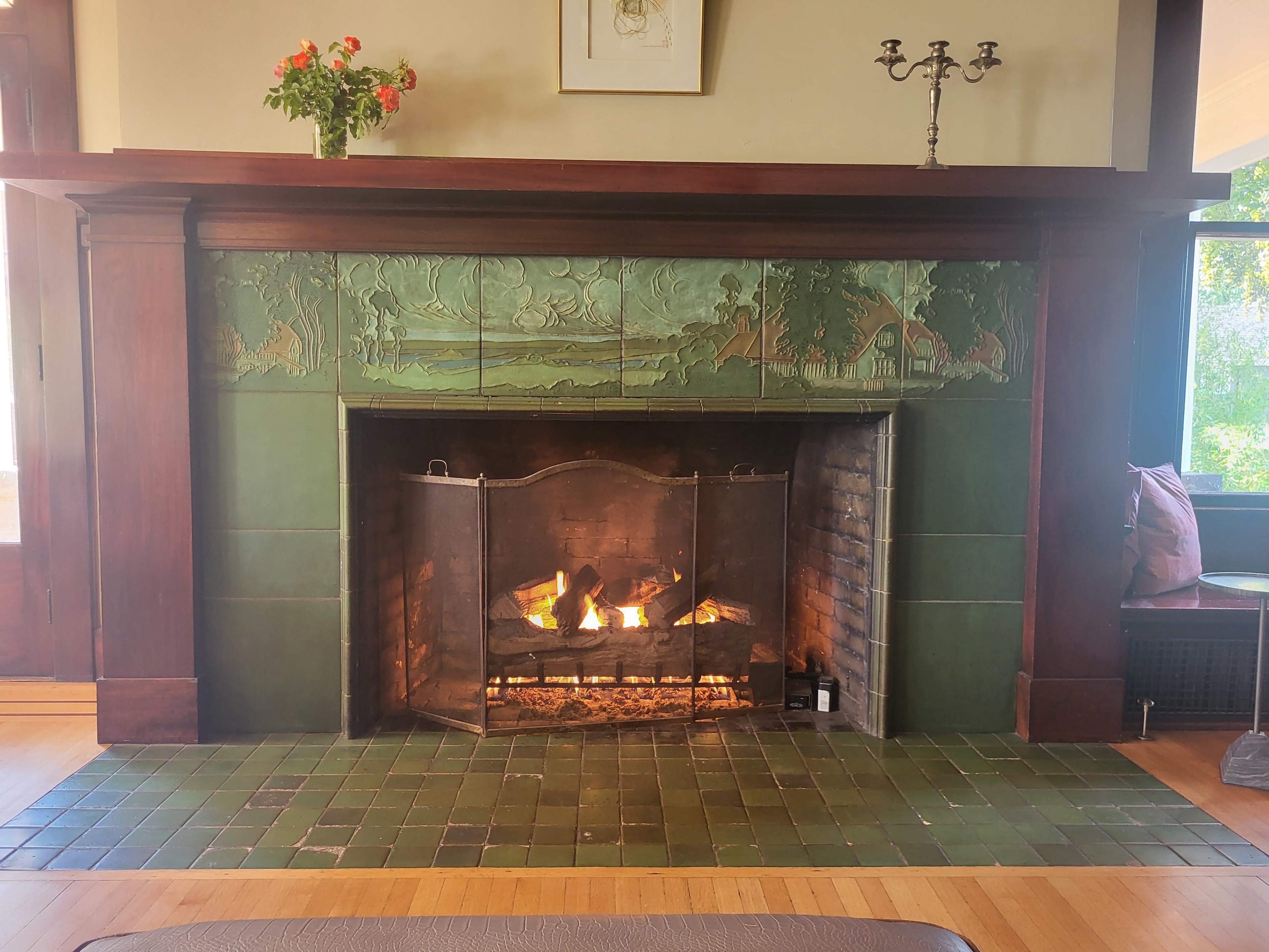 The image shows a fireplace with ornate green tiles and a wrought iron screen, featuring a fire burning within.