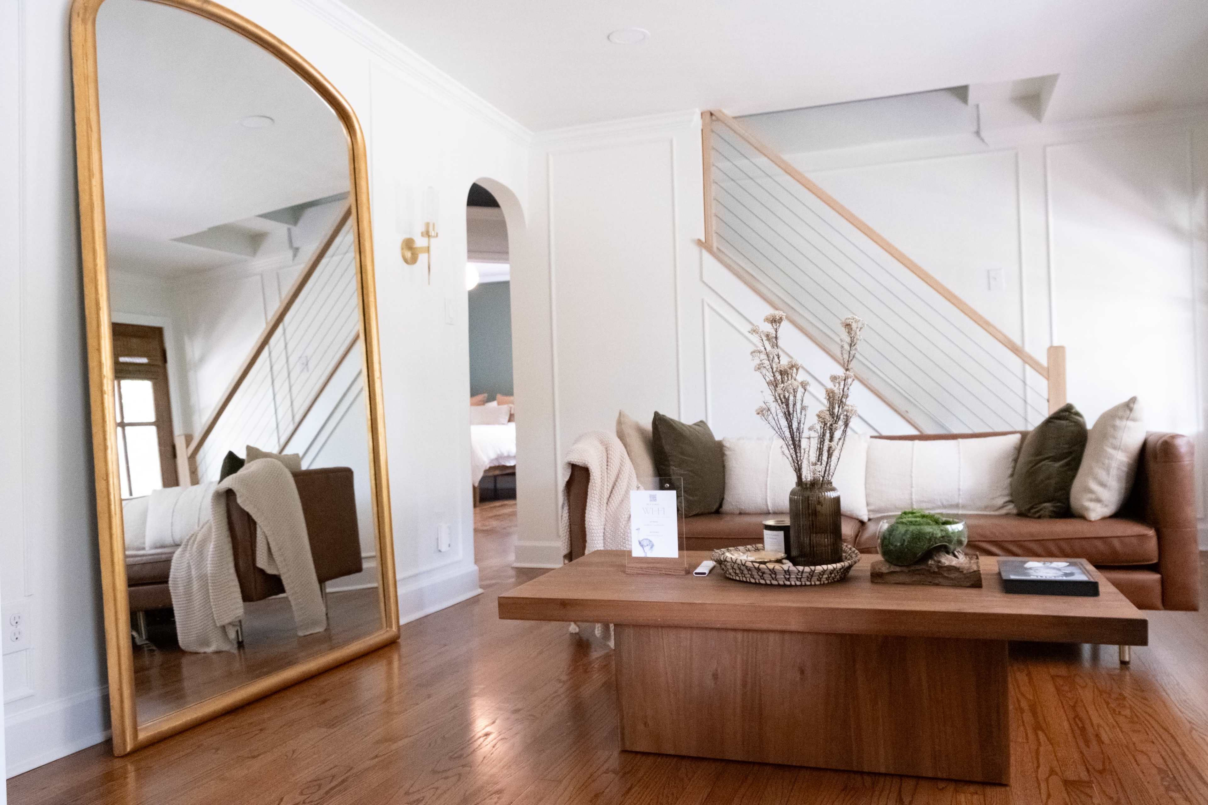 A well-decorated living room features a brown leather sofa, a wooden coffee table, and a large mirror reflecting a staircase.