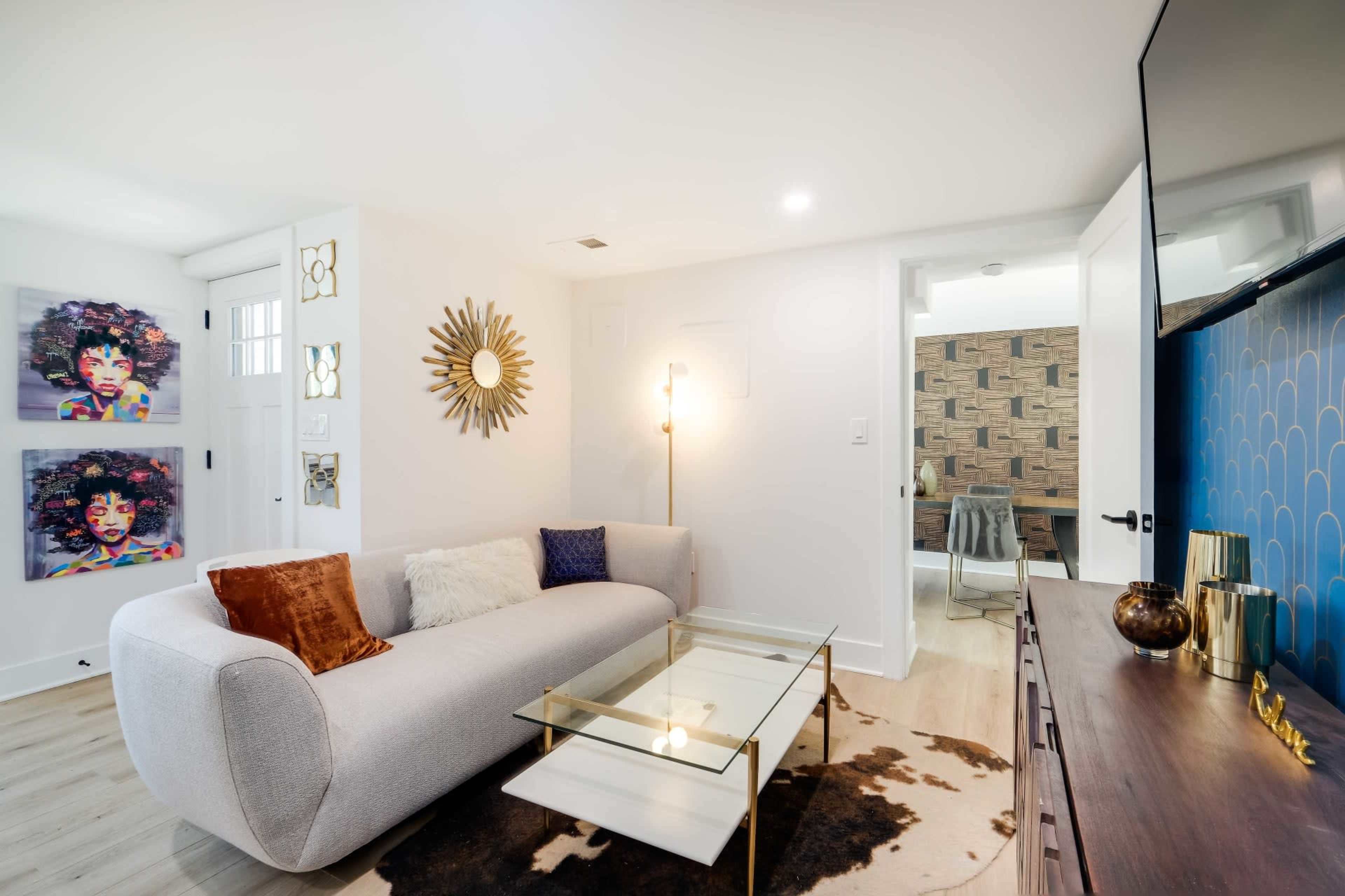 A modern living room with a curved gray sofa, a glass coffee table, and decorative artworks on the walls, including colorful portraits.