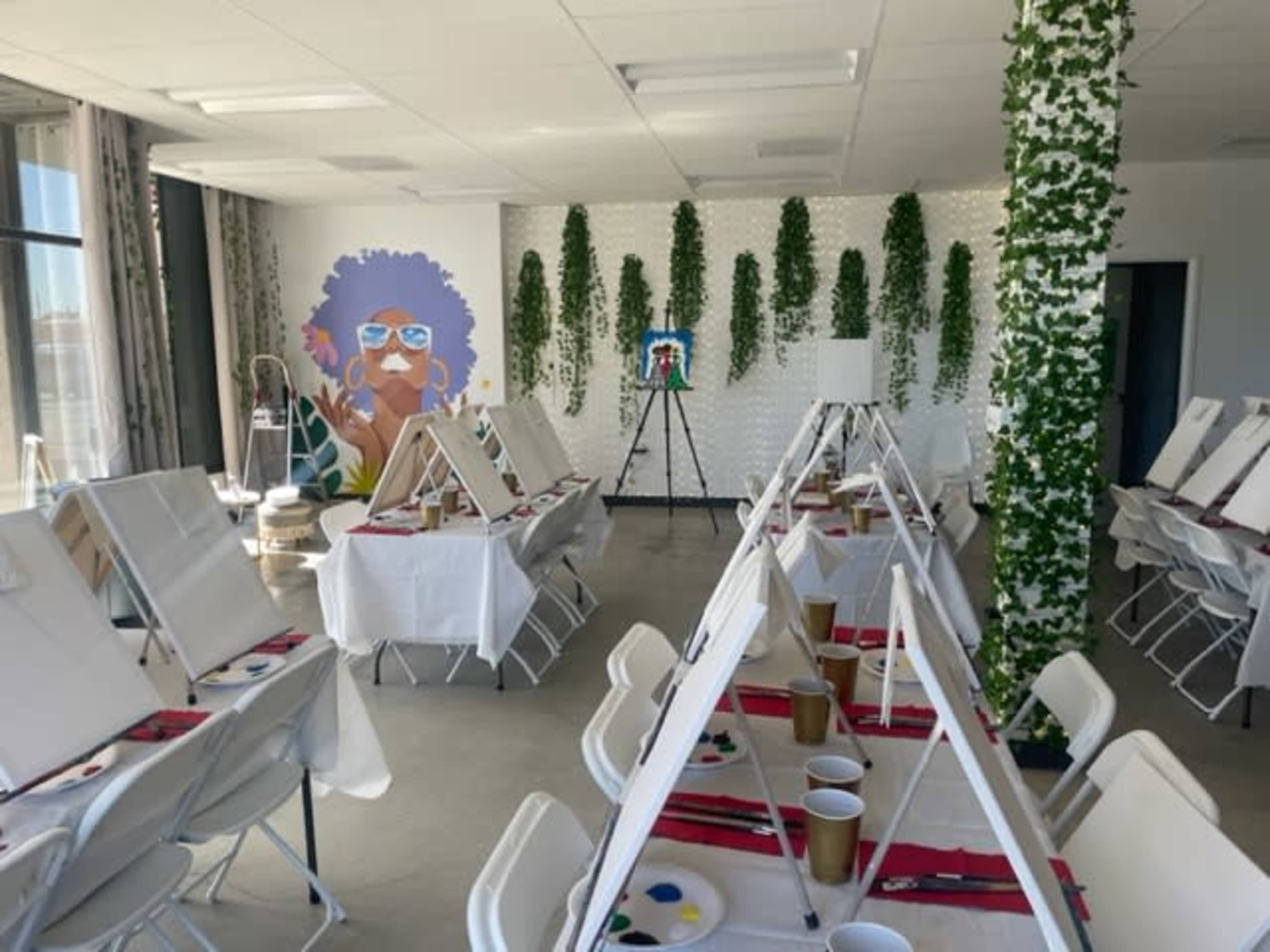 The image shows an art studio featuring easels with canvases, tables set with painting supplies, and a large mural of a woman with colorful hair on the wall.