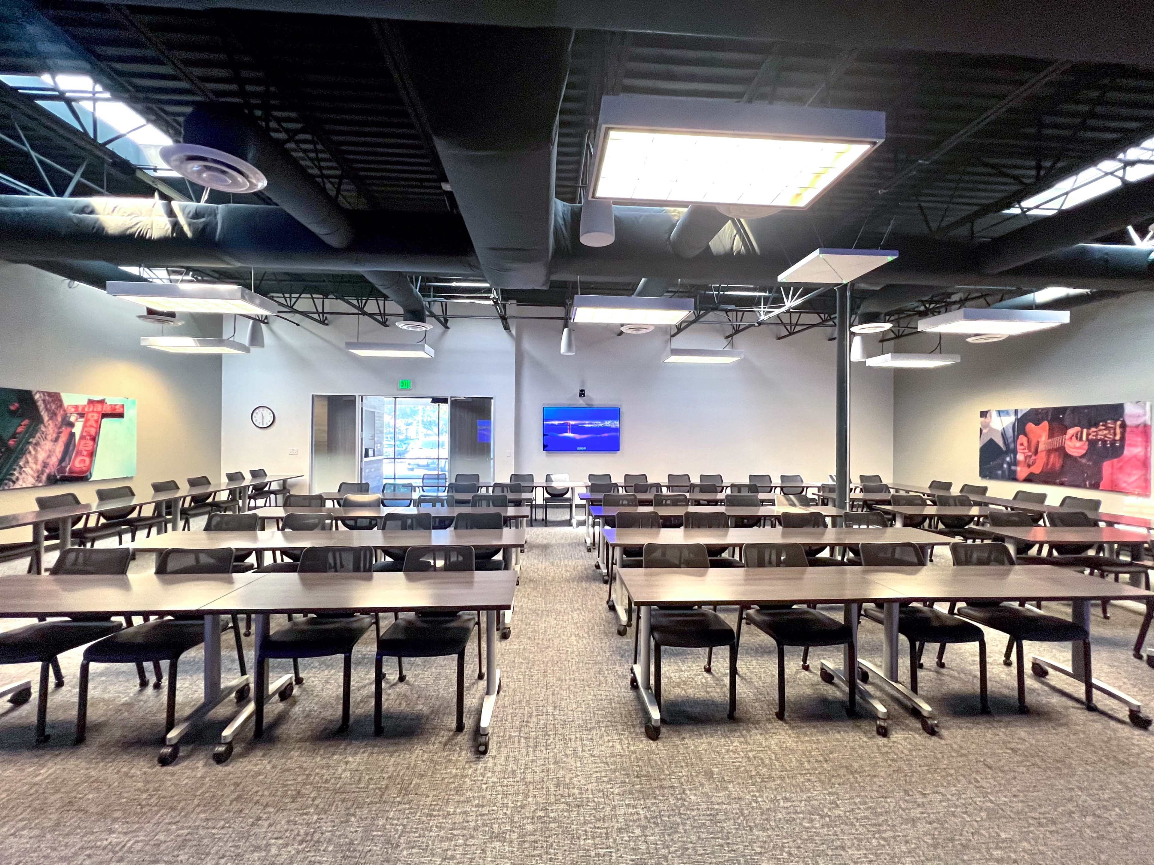 The image shows a modern classroom arranged with rows of tables and chairs, featuring a large screen at the front and vibrant wall graphics.