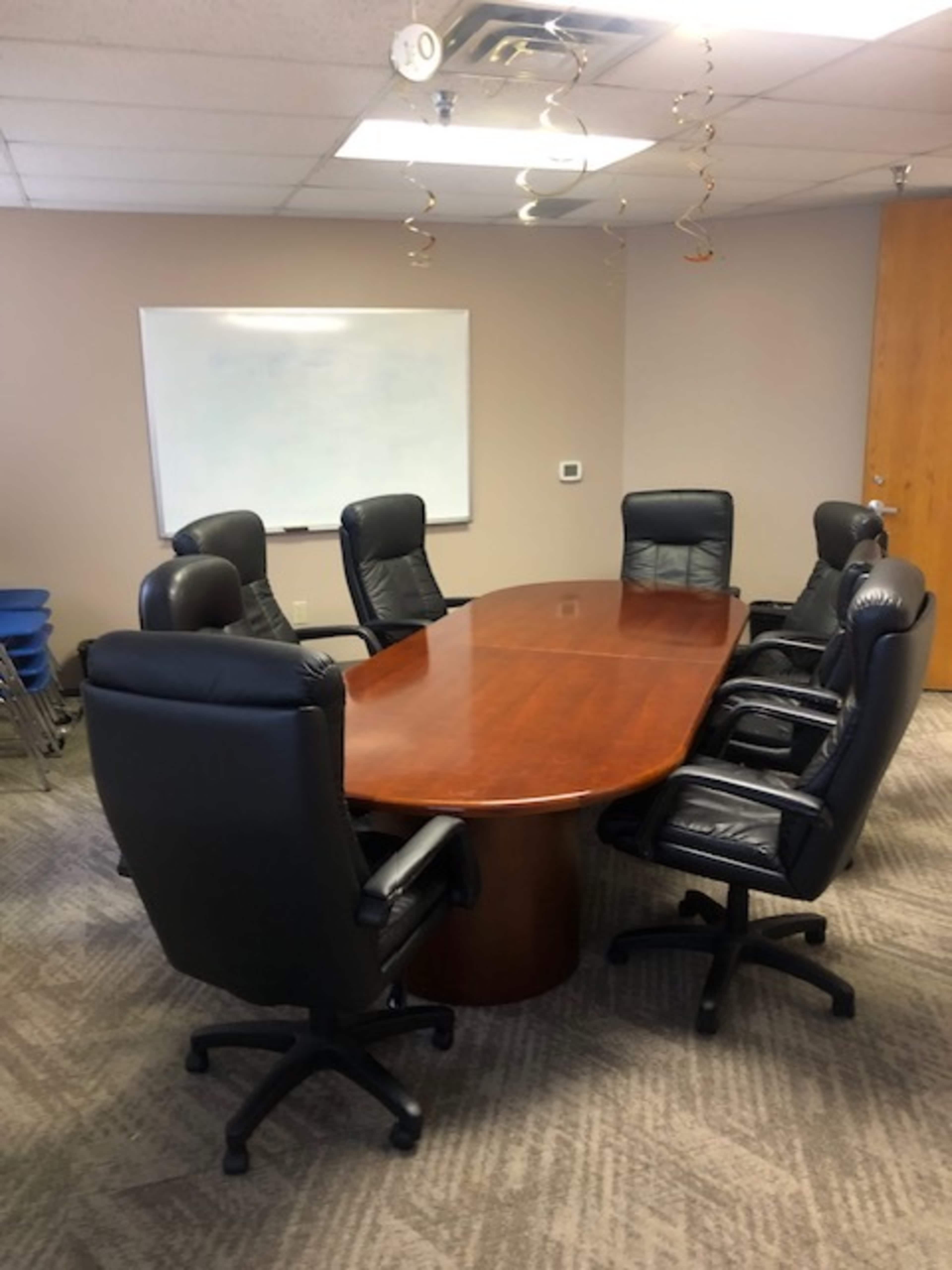 A conference room features an oval wooden table surrounded by eight black ergonomic chairs and a whiteboard on the wall.
