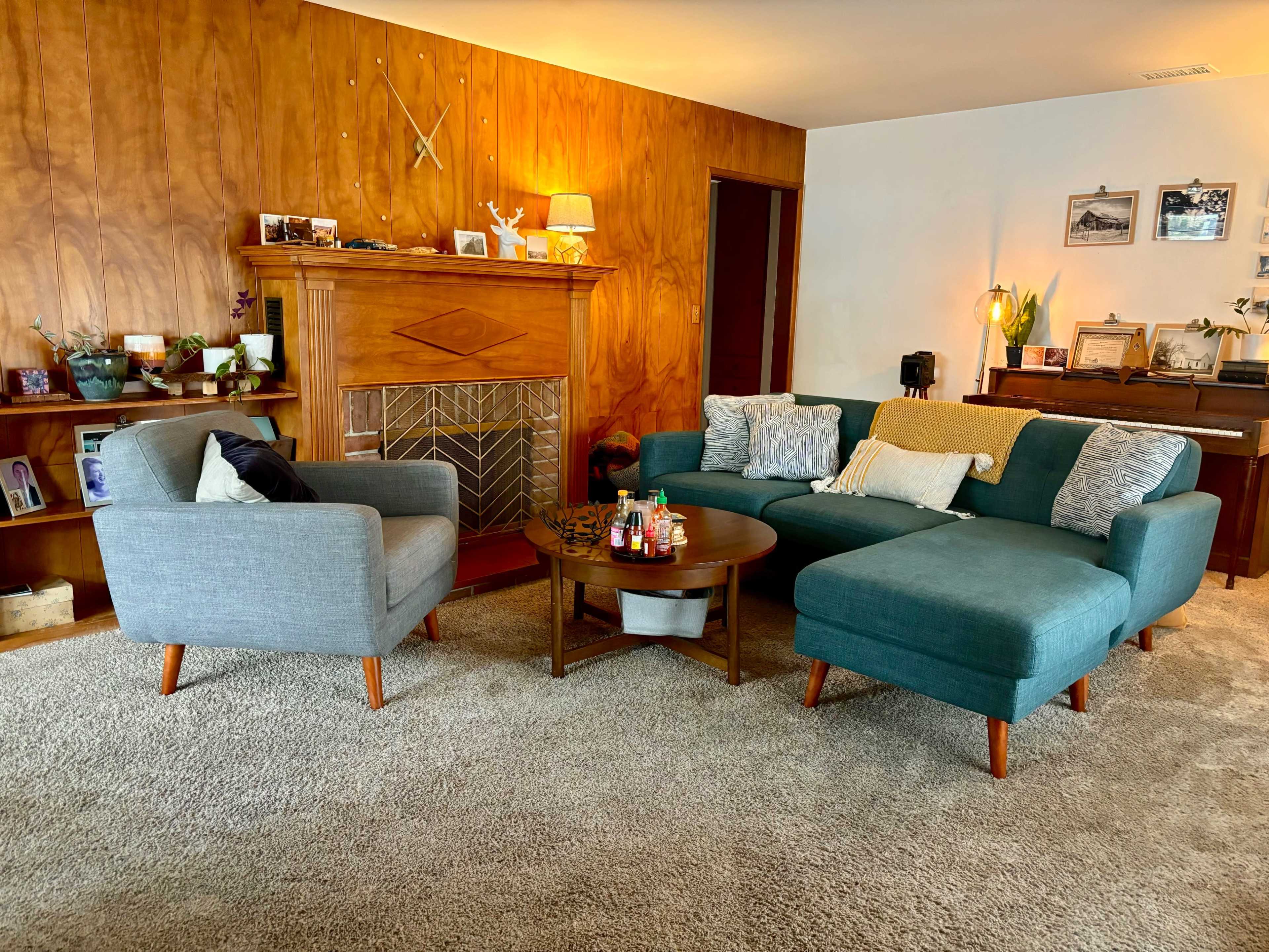 A cozy living room with a teal sectional sofa, a gray armchair, a wooden coffee table, and a fireplace adorned with decorative items against a wood-paneled wall.