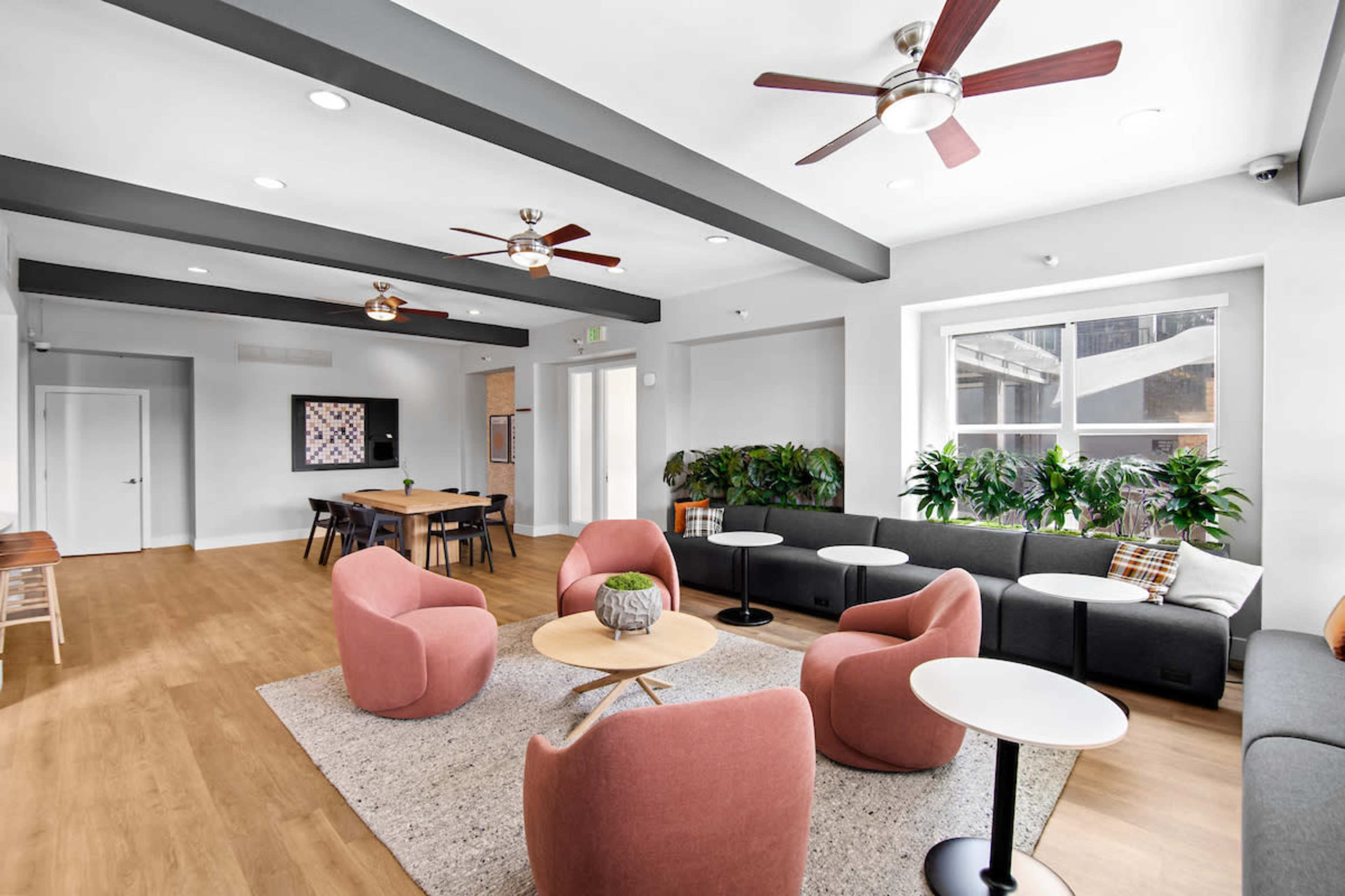 The image shows a modern common area featuring pink chairs, a gray sofa, and a light wood floor, with plants and ceiling fans adding to the decor.