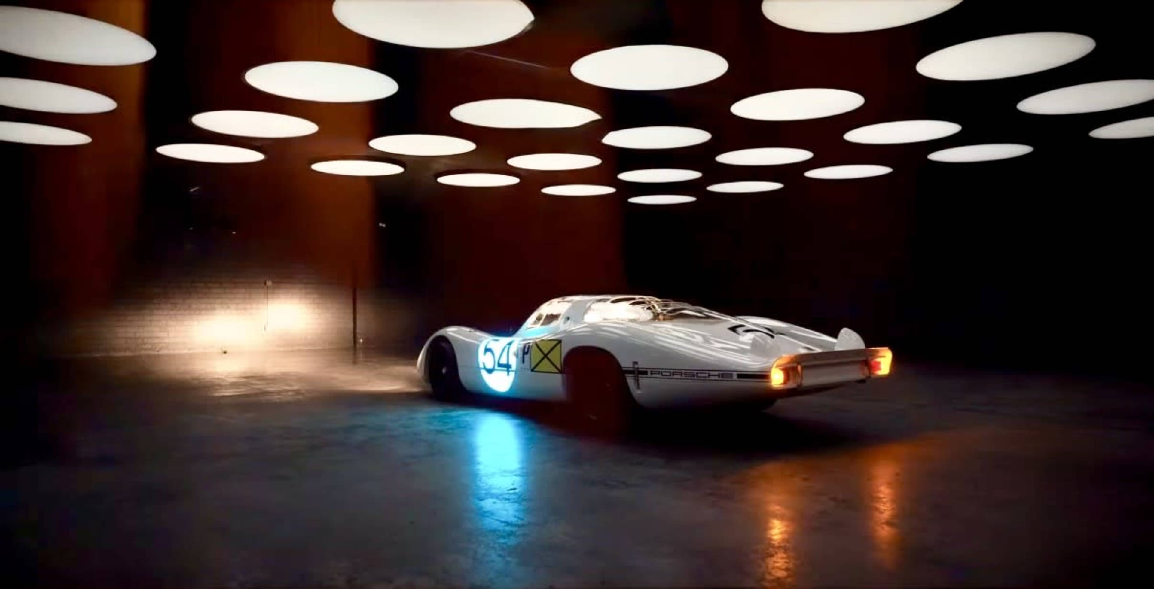 A vintage Porsche sports car is positioned in a dimly lit room with circular overhead lights illuminating its sleek body.