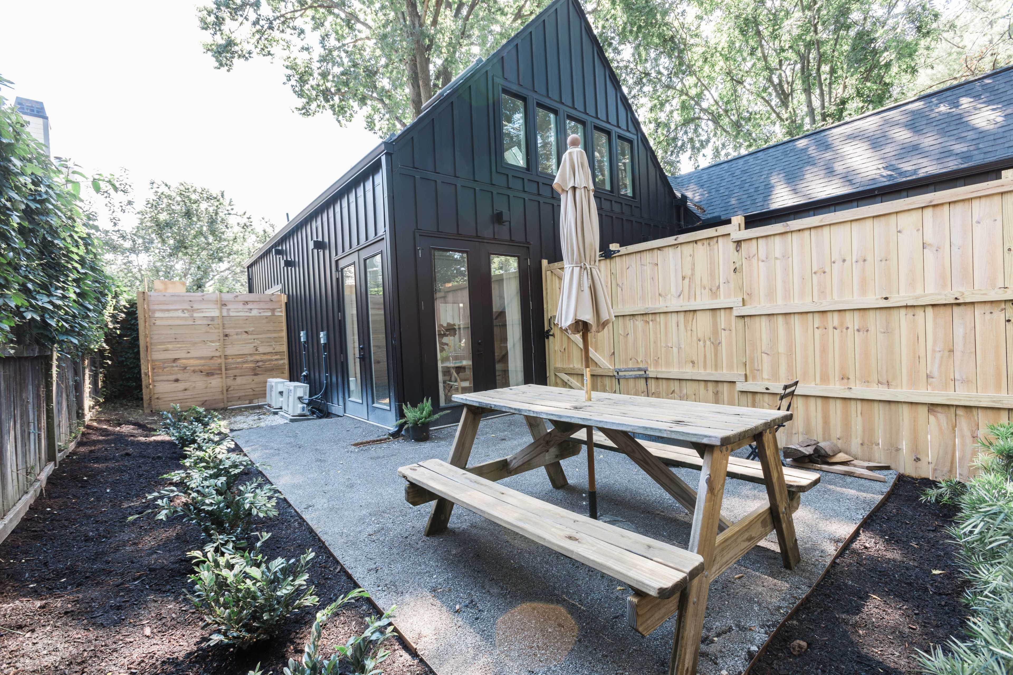 A modern black-paneled house stands behind a wooden picnic table in a gravel patio, surrounded by a wooden fence and landscaped with greenery.