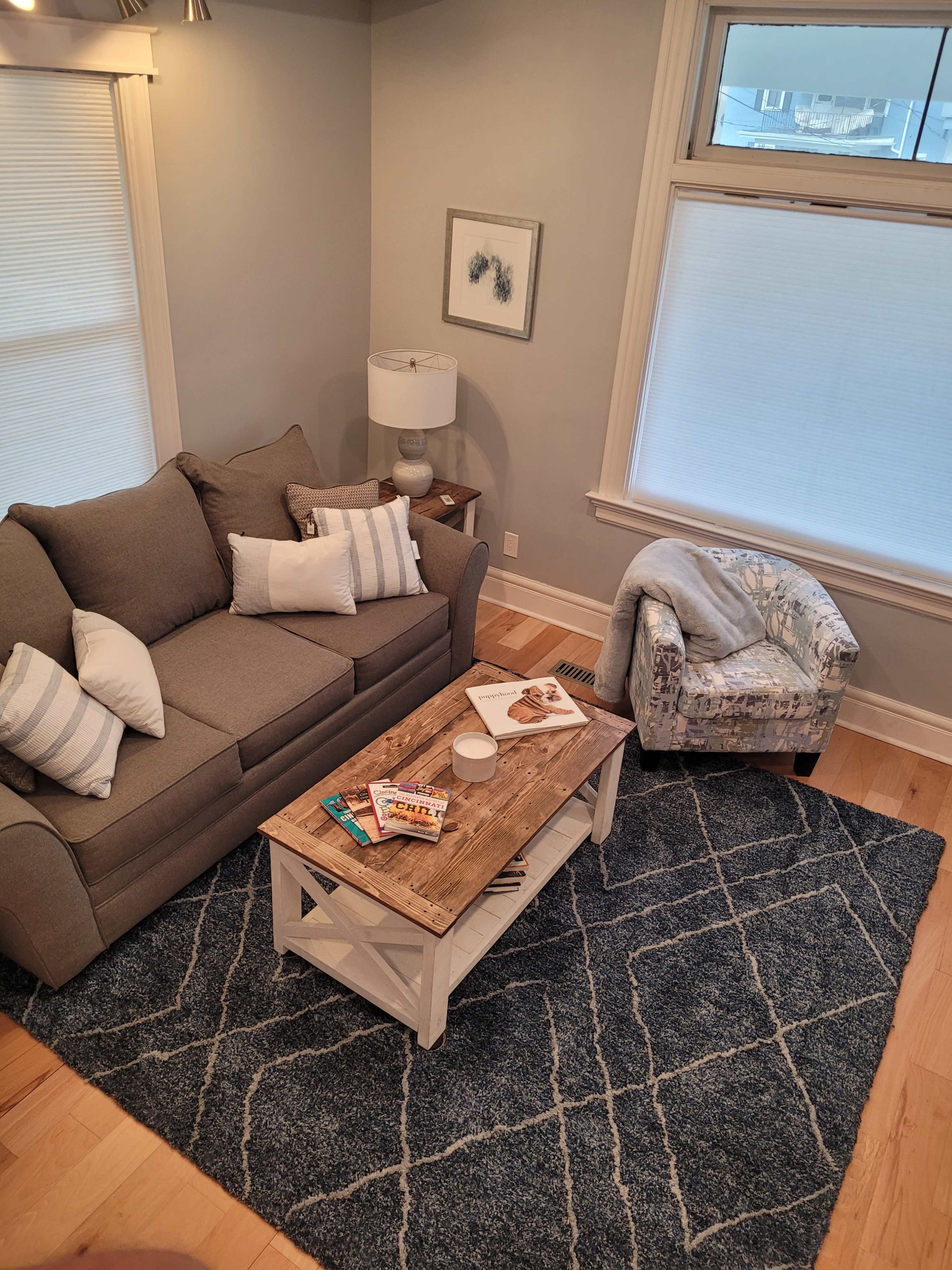 A cozy living room with a grey couch, a wooden coffee table topped with magazines, and a patterned armchair on a blue area rug.