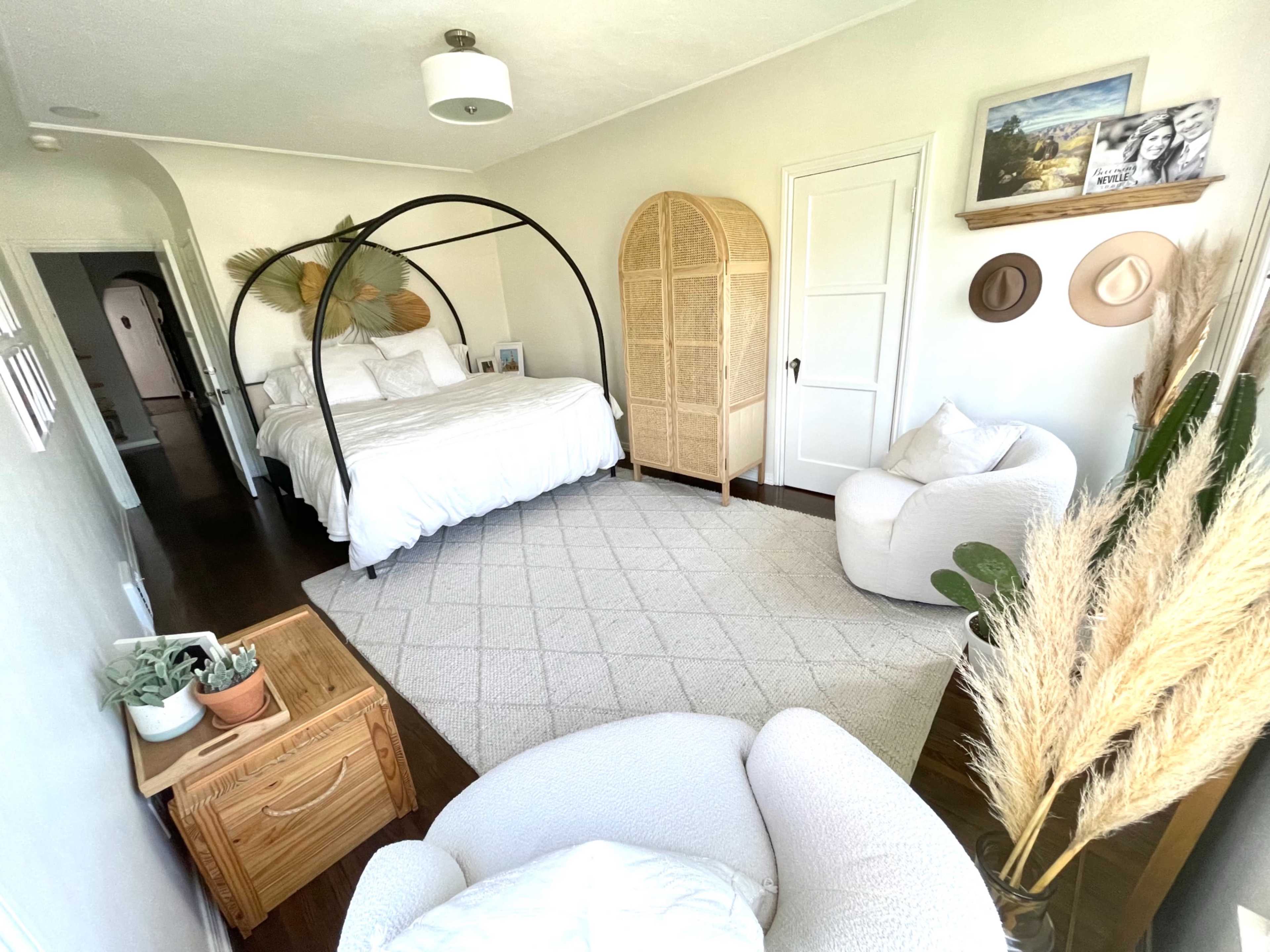A bright bedroom features a large bed with a black metal frame, wicker accents, a cozy chair, and decorative elements like a plant and framed photos.