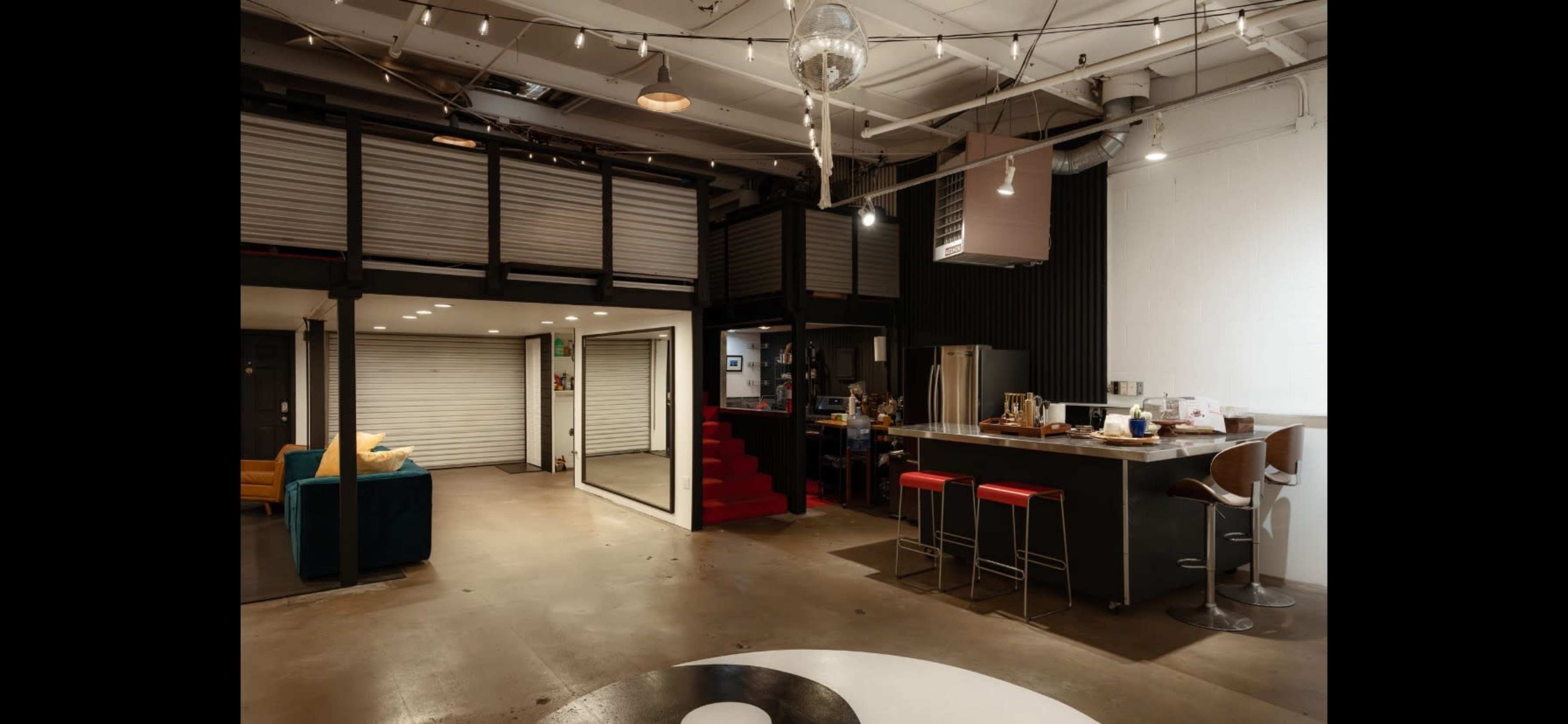 An industrial-style open-concept living space featuring a kitchen bar with red stools, a living area with a green couch, and a lofted section accessible by stairs.