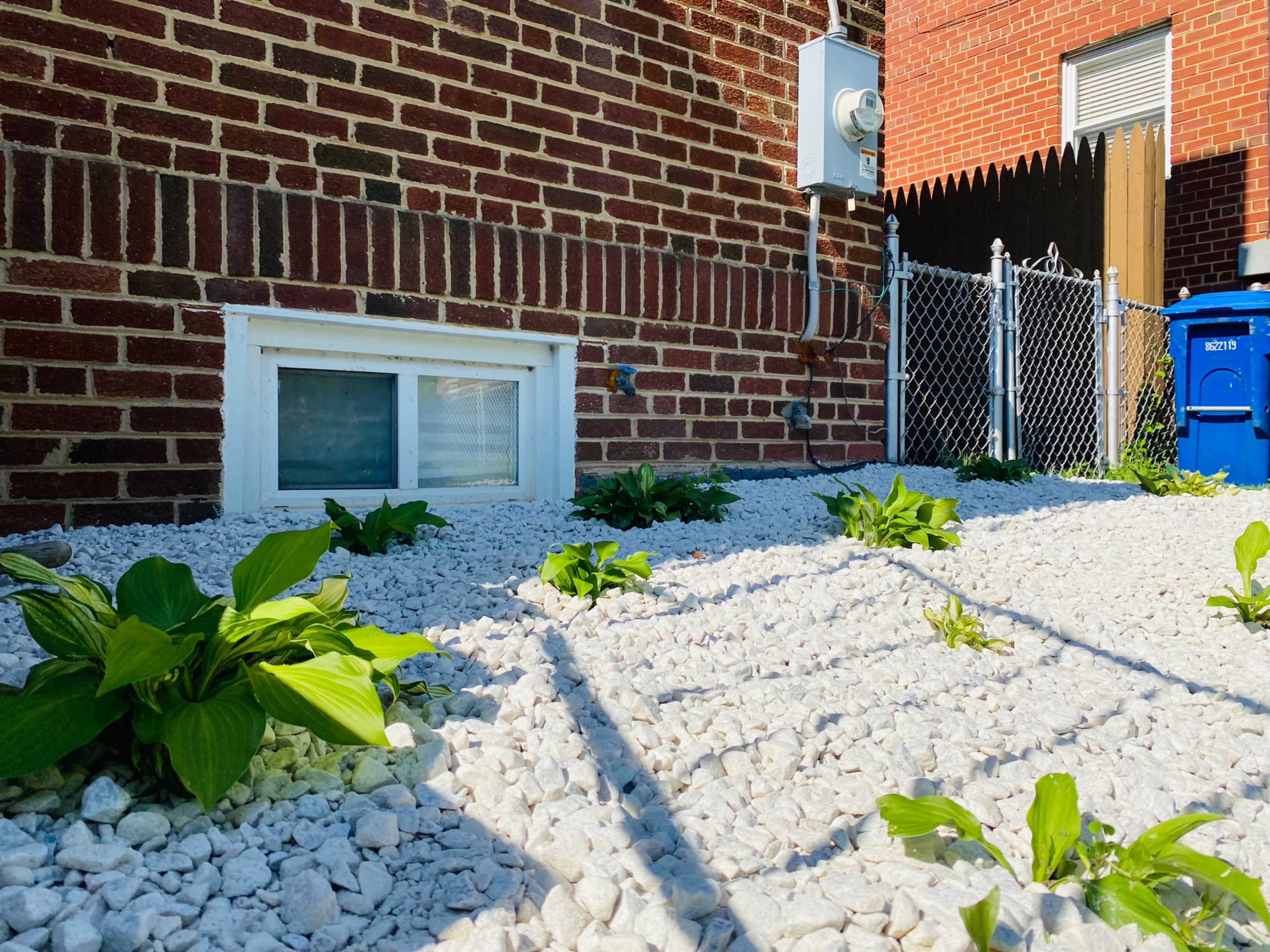 The image shows a small garden area with white gravel surrounding green plants next to a brick wall and a fenced section with a blue trash bin.