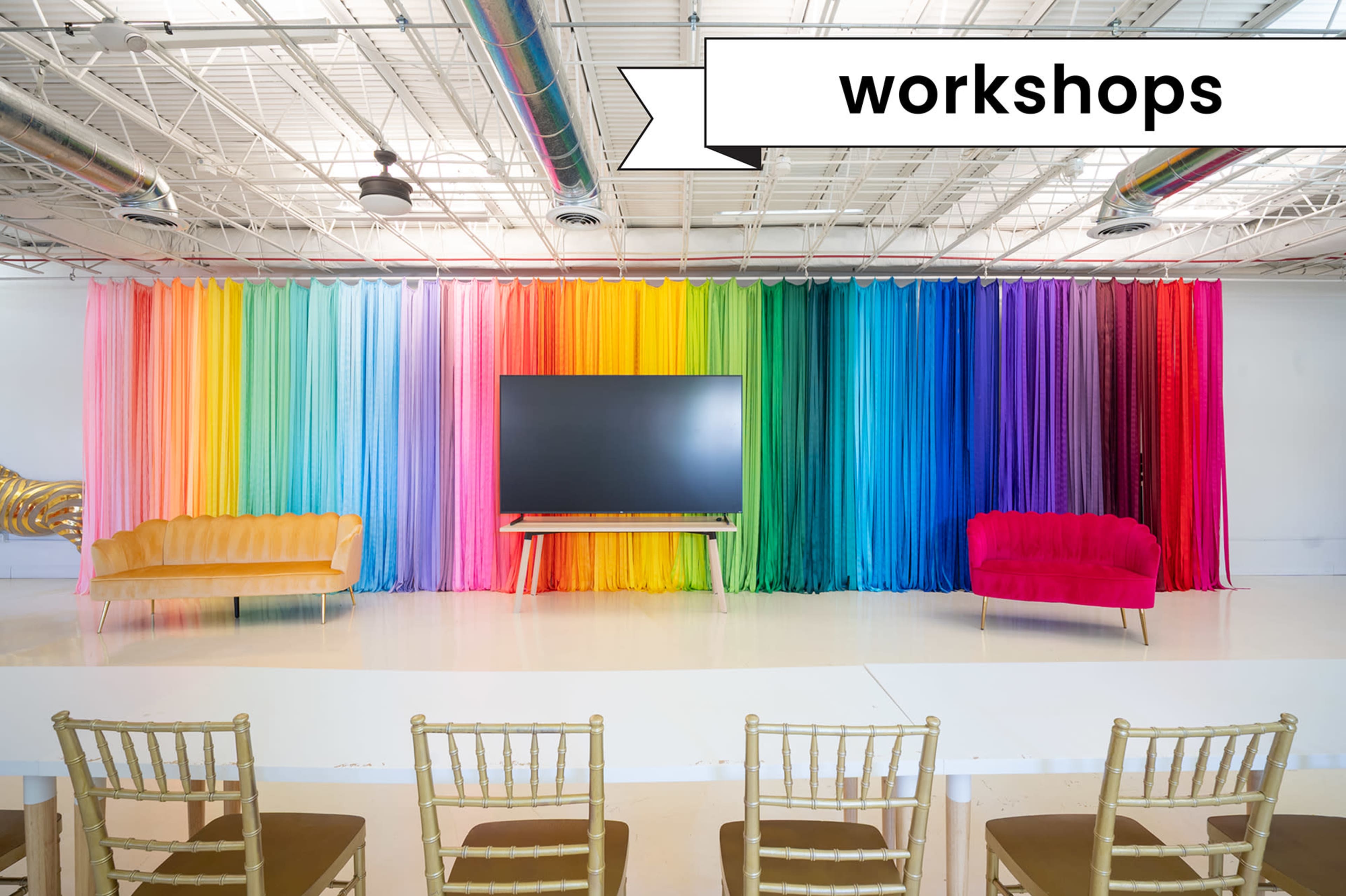 A brightly colored fabric backdrop is arranged in a rainbow gradient behind a large television, with two sofas placed in front and a long table with gold chairs in the foreground.