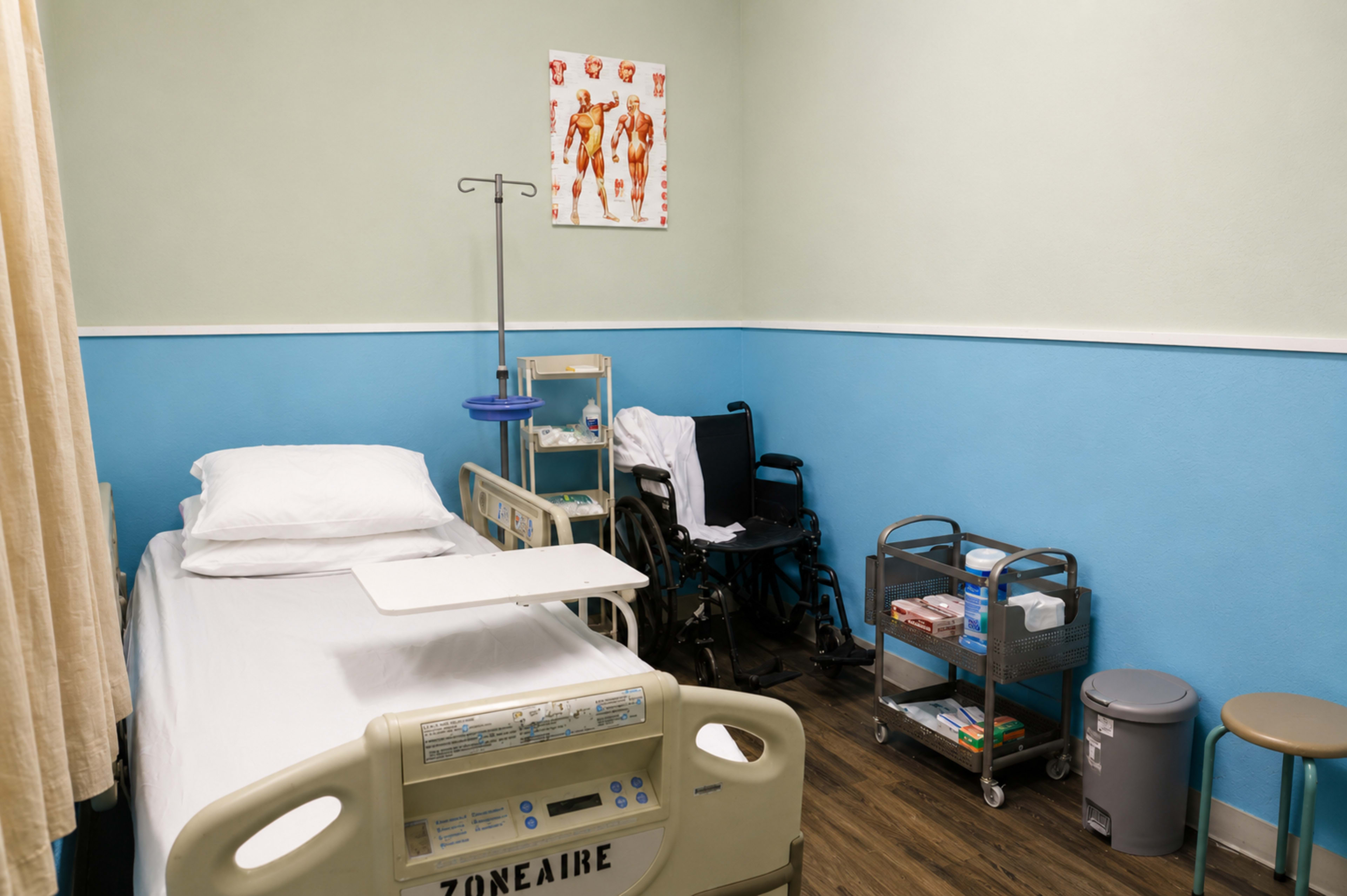 The image shows a small hospital room with a bed, a wheelchair, and medical supplies organized on a cart.