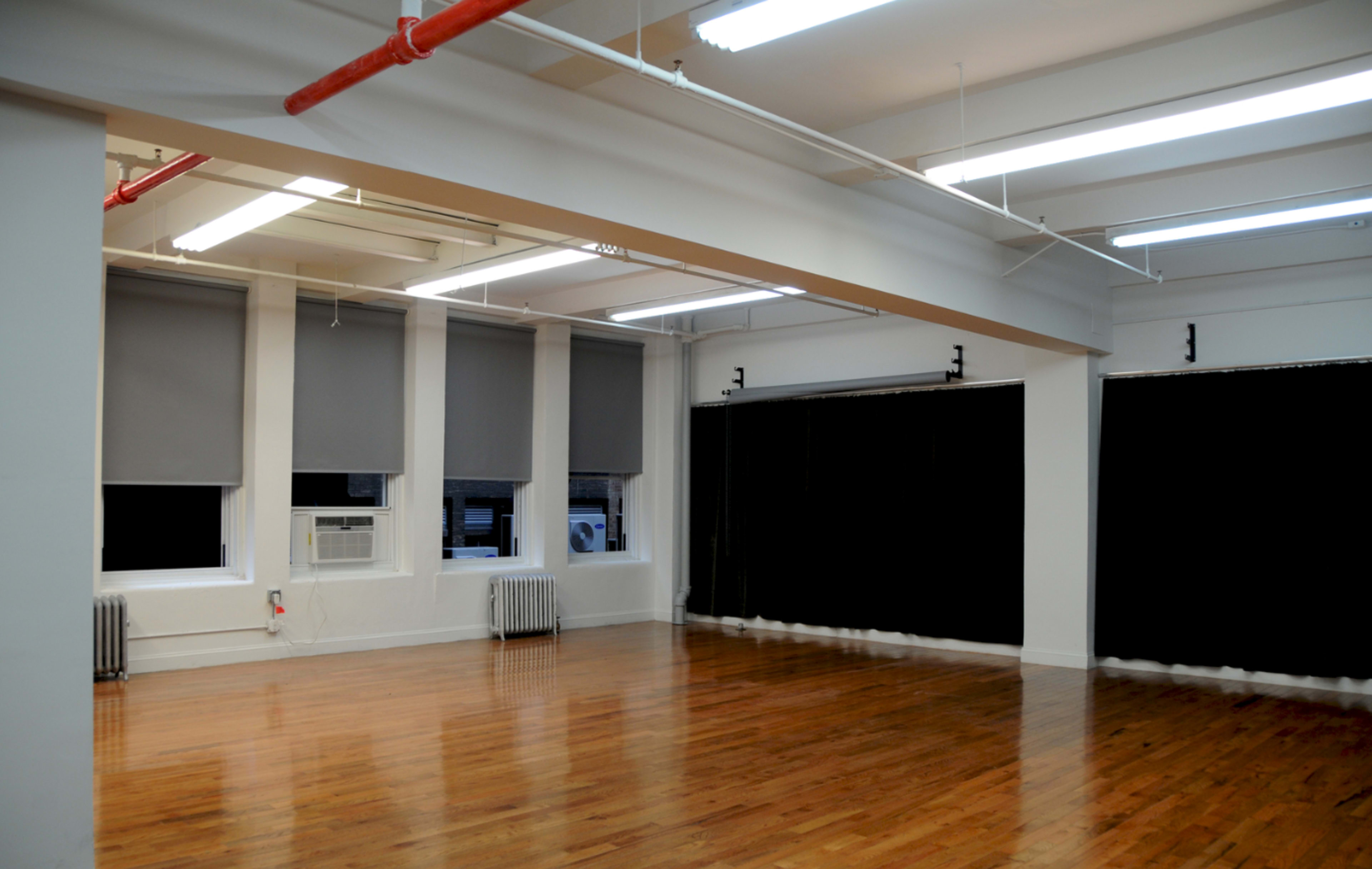 A spacious, bright room with hardwood floors, large windows covered by gray blinds, and an air conditioning unit.