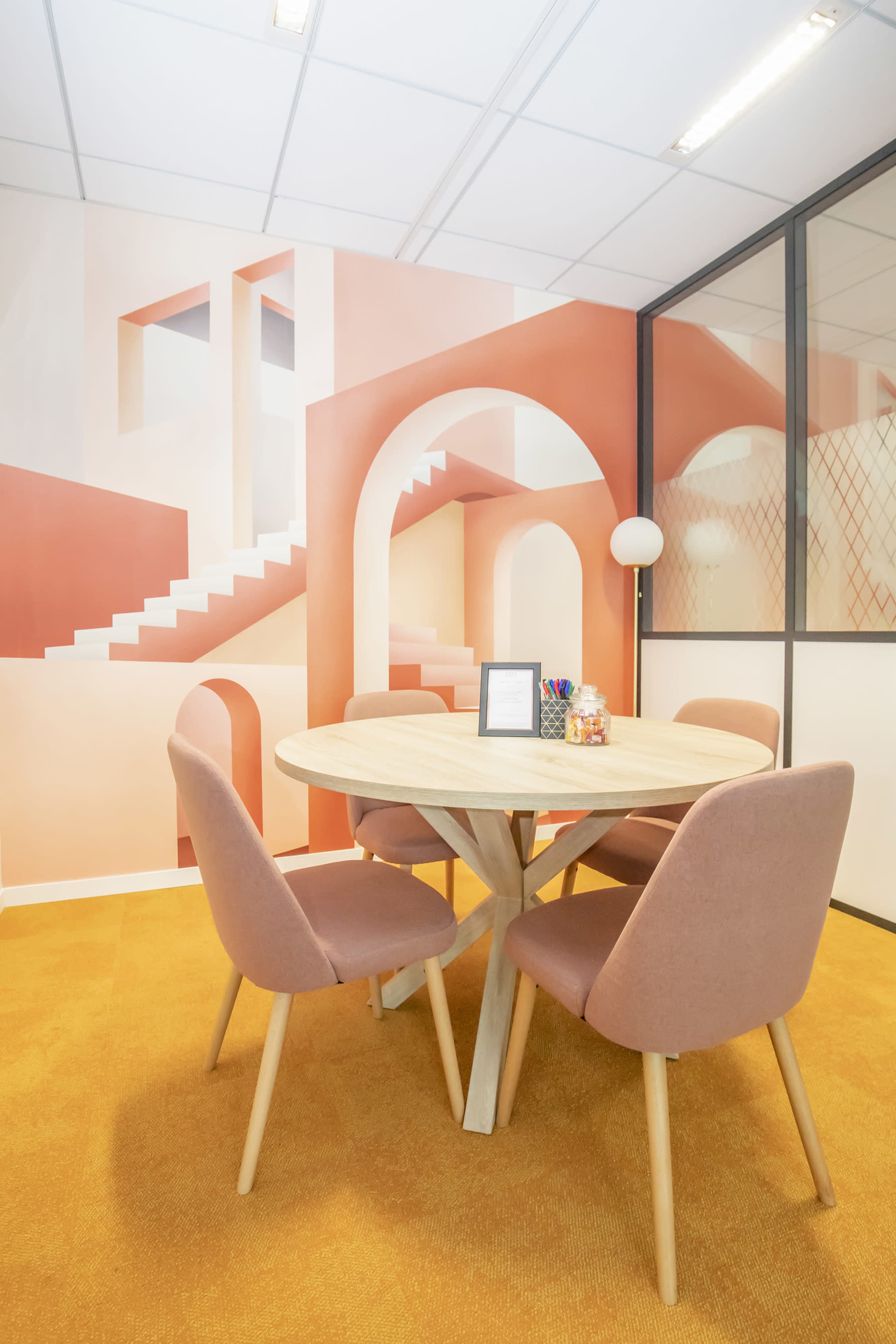 A modern office space features a round wooden table surrounded by four upholstered chairs, with a colorful mural of abstract architectural shapes on the wall.