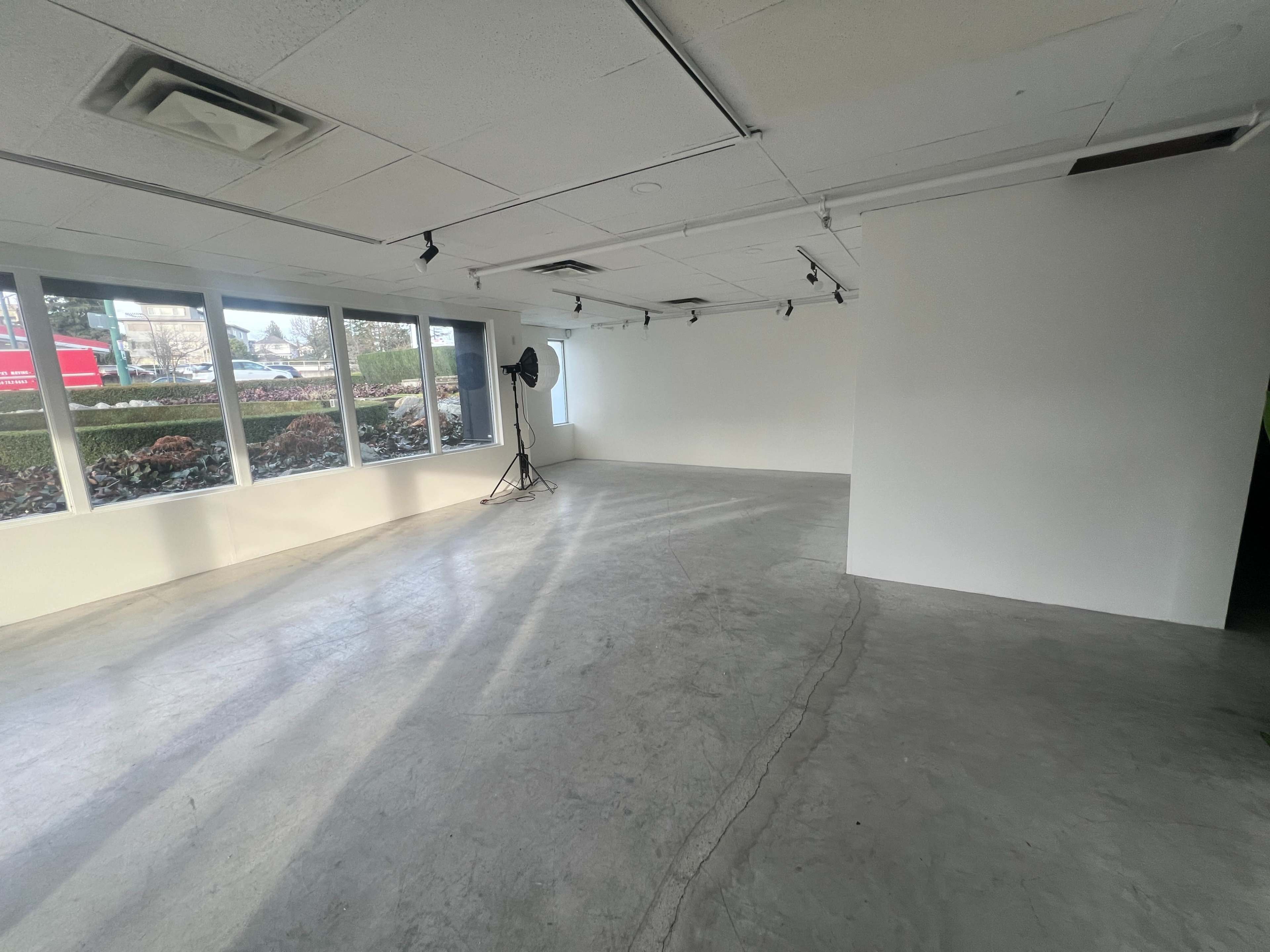 The image shows a spacious, empty room with concrete flooring, large windows, and a backdrop stand set up against the wall.