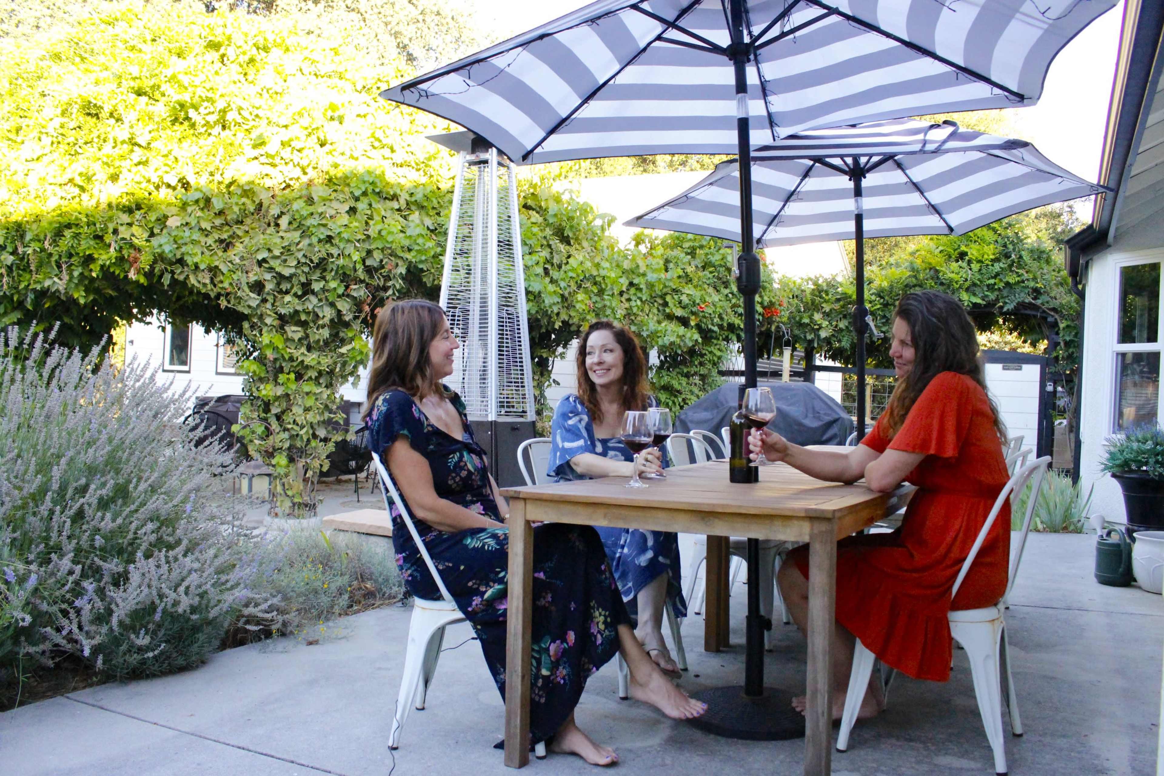 Three women sit around a wooden table under striped umbrellas in a garden setting.