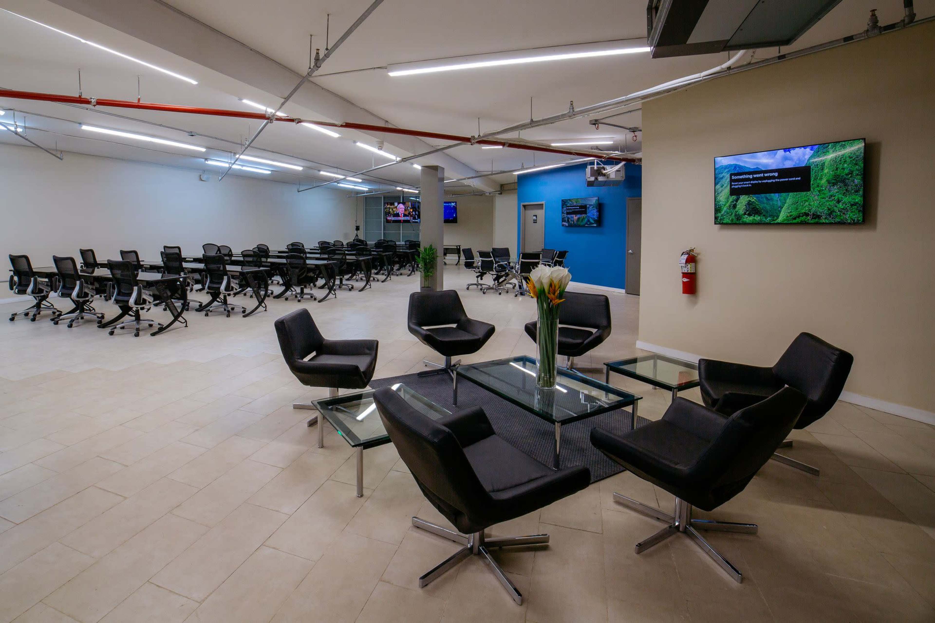 The image shows a modern office space with a seating area featuring black chairs around a glass table, and a large room in the background filled with empty office chairs and screens on the walls.