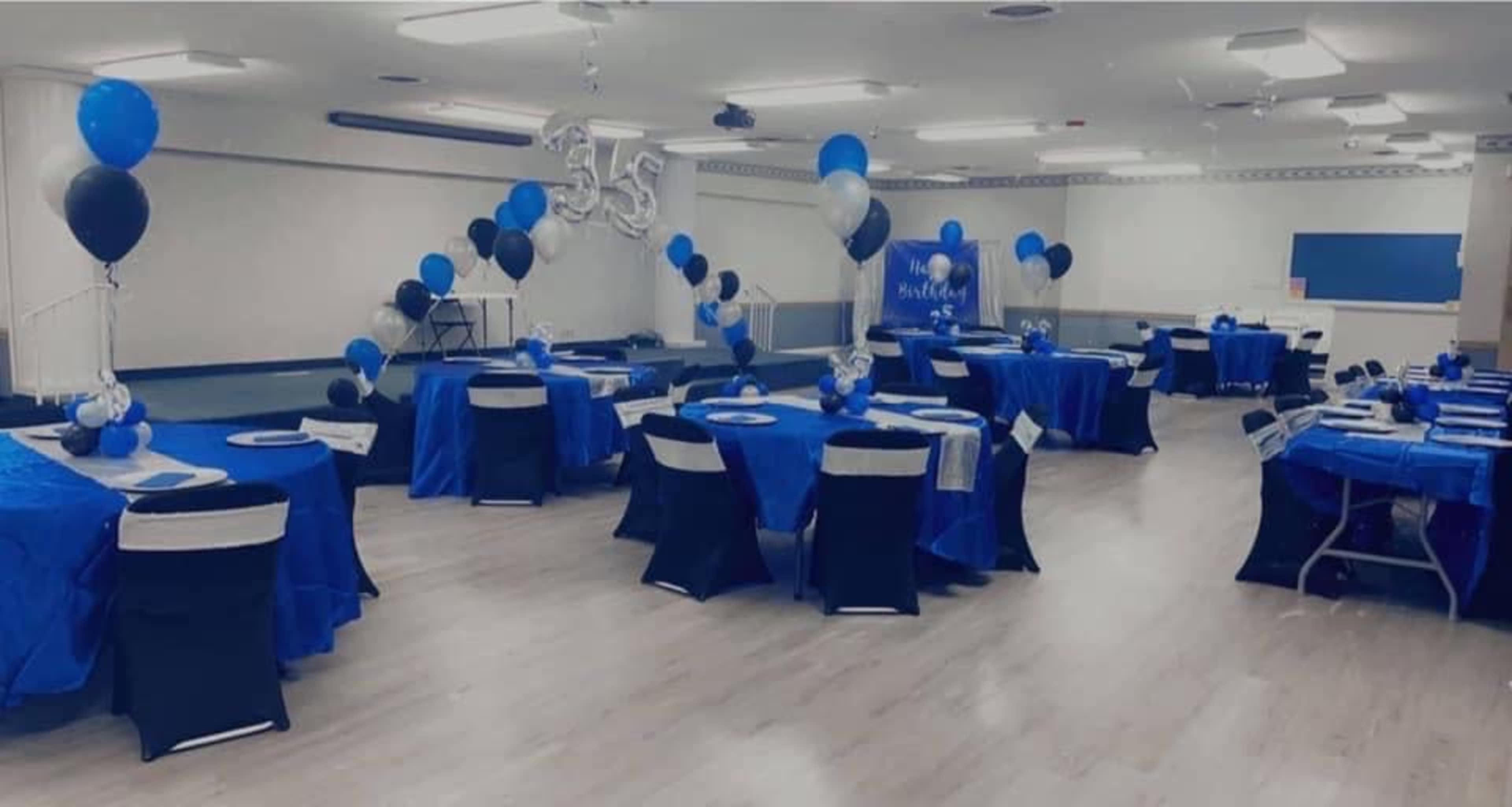 A decorated event space with tables set in blue and black, featuring balloons and a banner for a celebration.