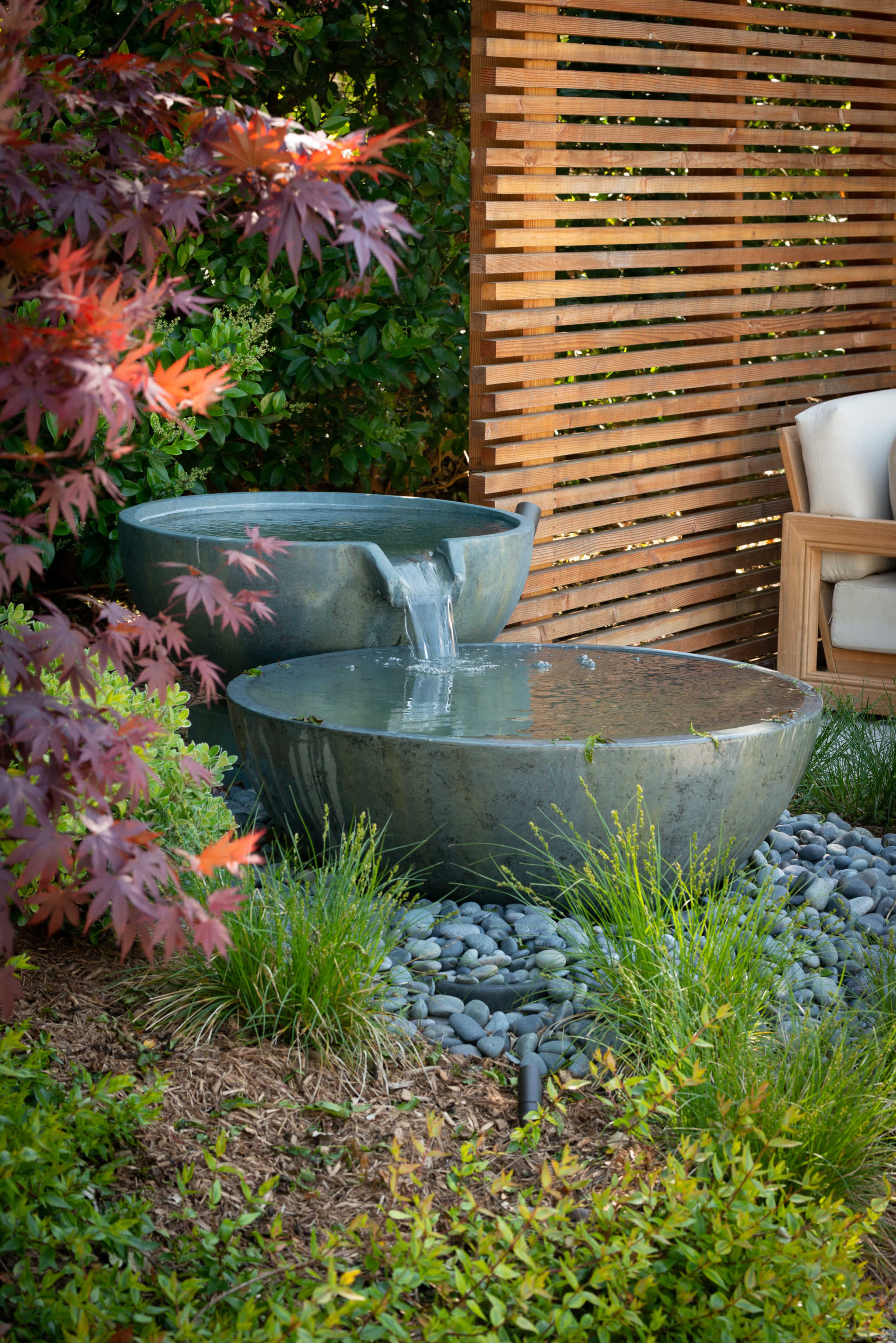 A water feature consisting of two stone basins, one pouring into the other, surrounded by decorative plants and rocks, set against a wooden slatted backdrop.