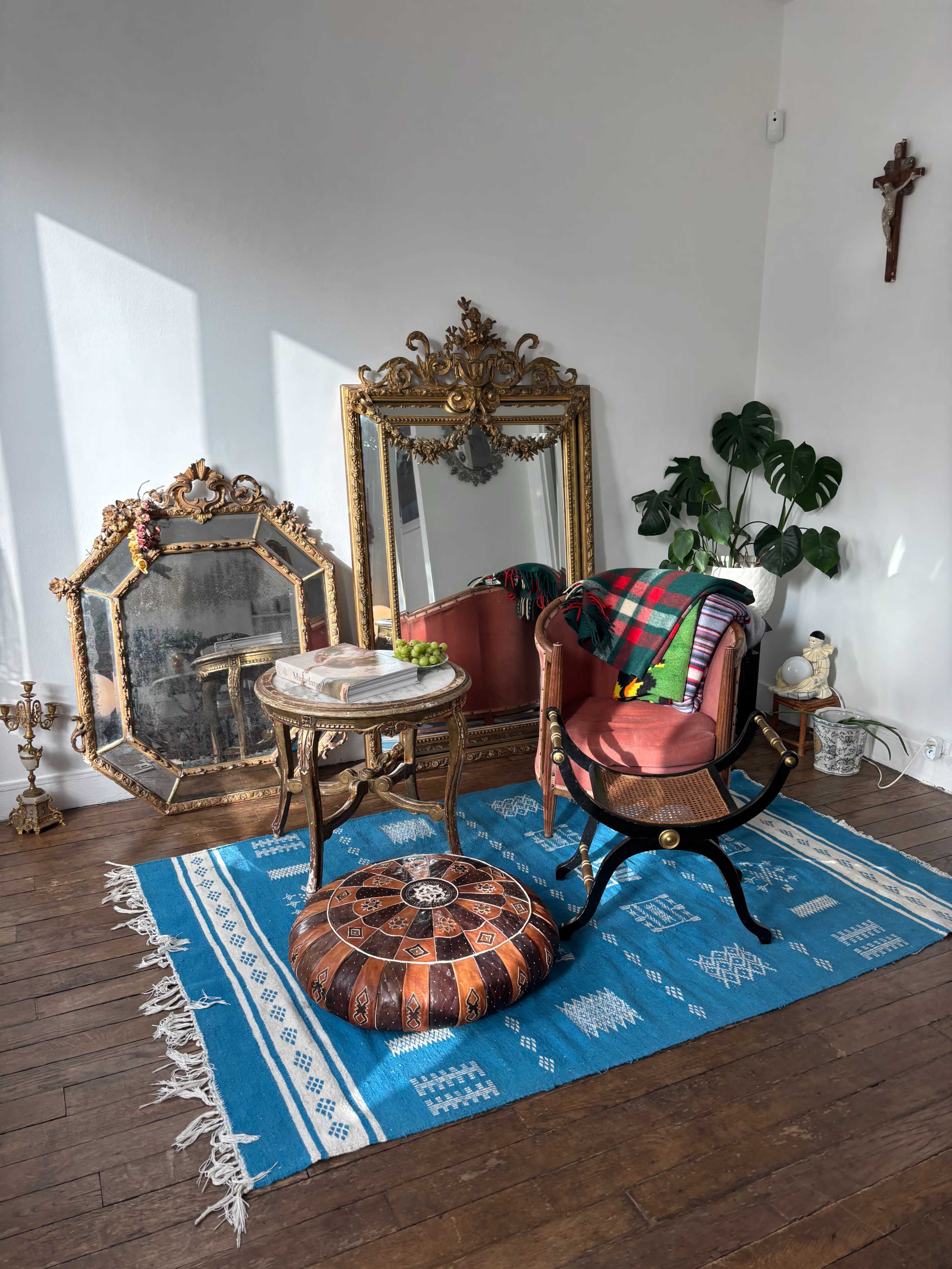 The image shows a cozy corner featuring a vintage mirror, an ornate chair with a colorful blanket, a decorative ottoman, and a potted plant on a blue patterned rug.