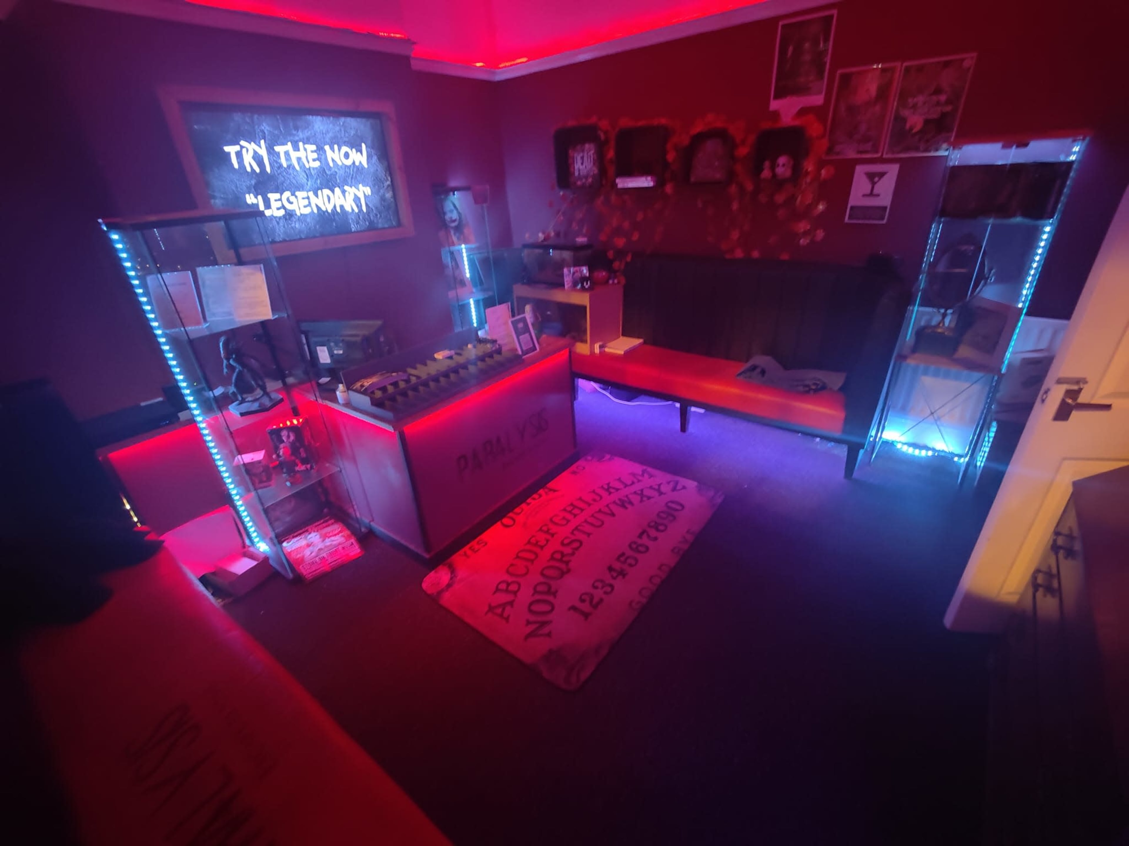 The room features red walls with neon lighting, two benches, a central counter with an ouija board design, and a large illuminated sign that reads "TRY THE NOW #LEGENDARY."