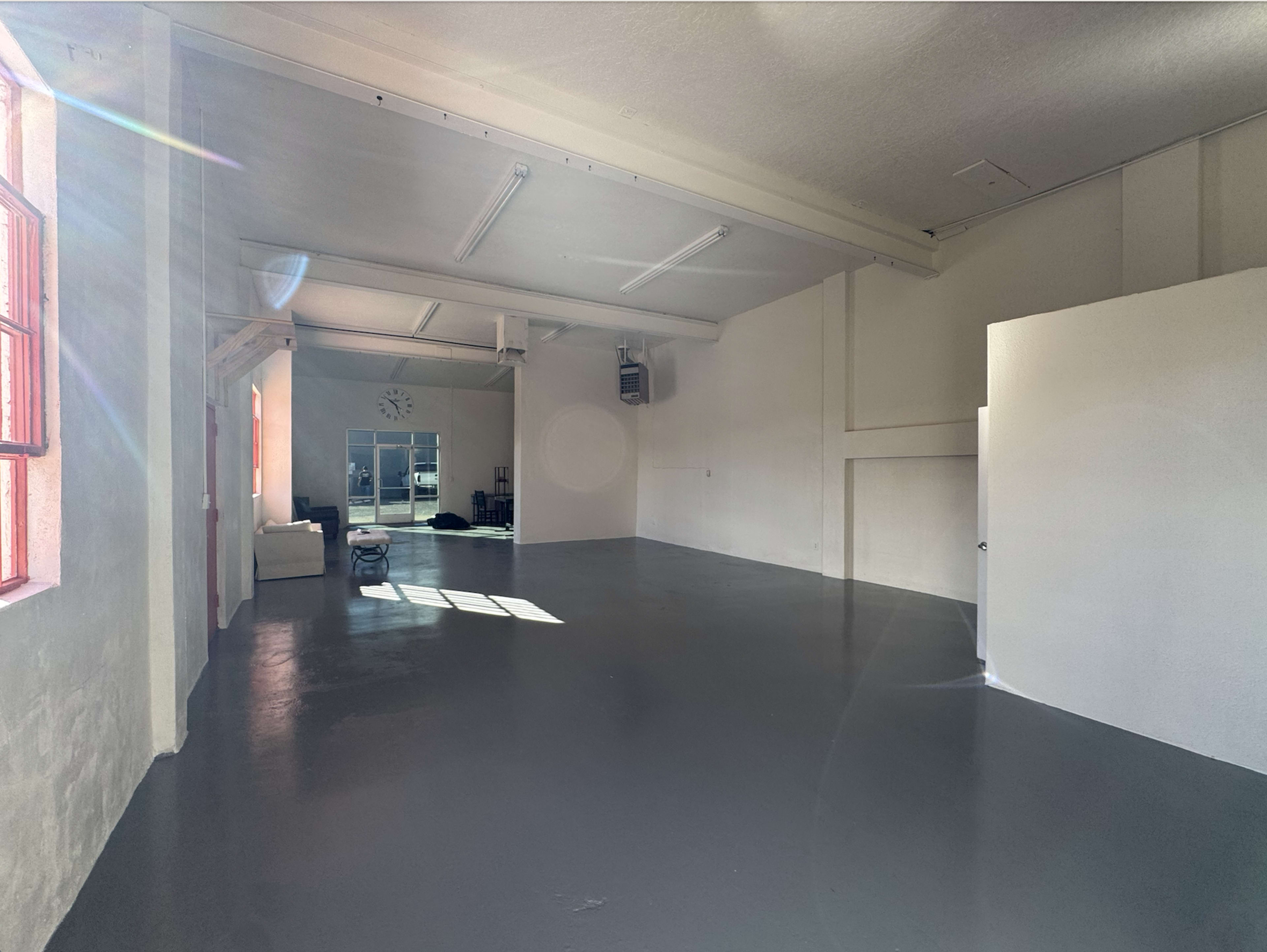 The image shows a spacious, empty industrial-style room with large windows, concrete flooring, and minimal furniture.