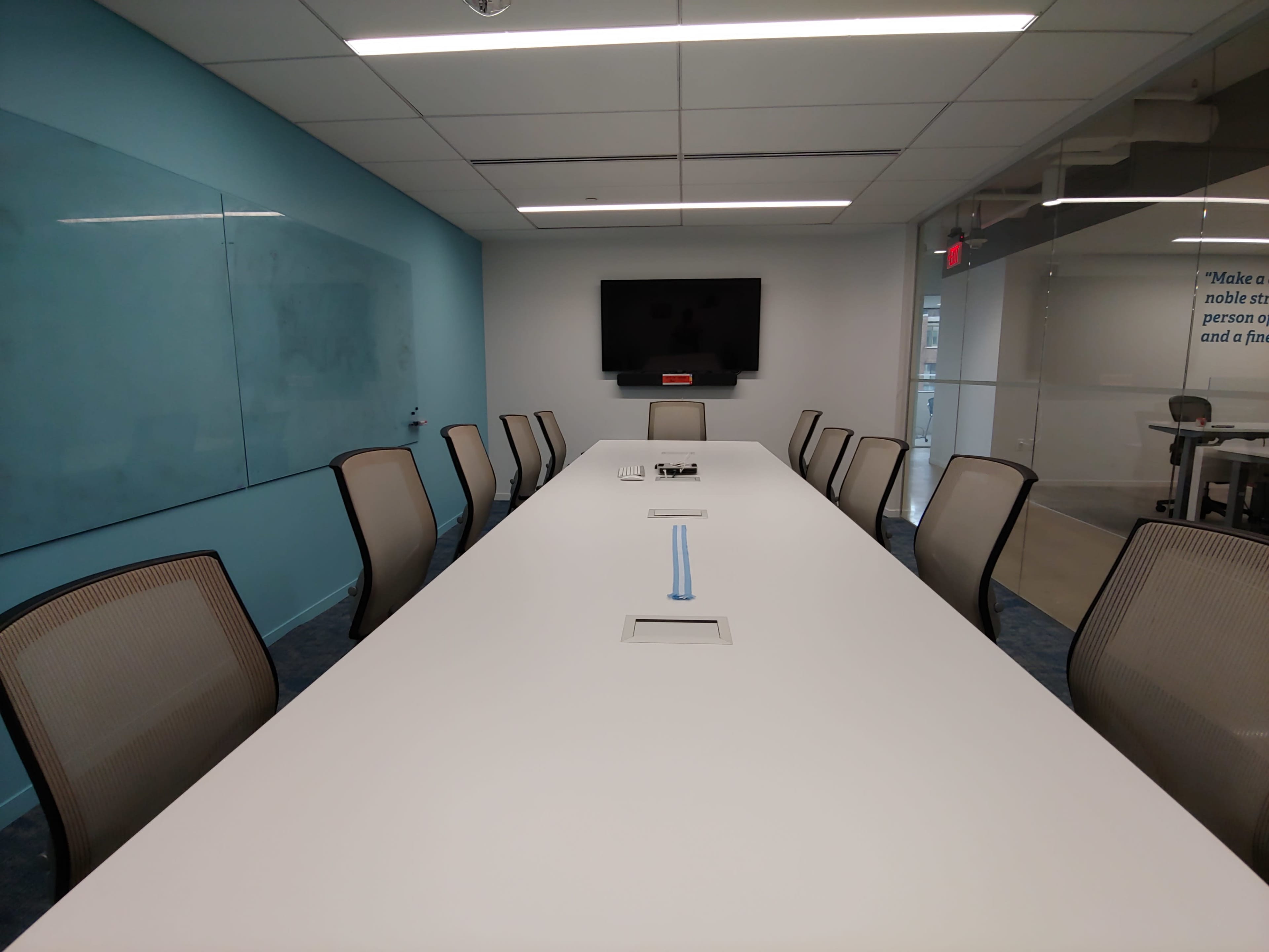 A long, modern conference table with multiple chairs surrounds it, positioned in a well-lit meeting room featuring a wall-mounted television and a glass partition.