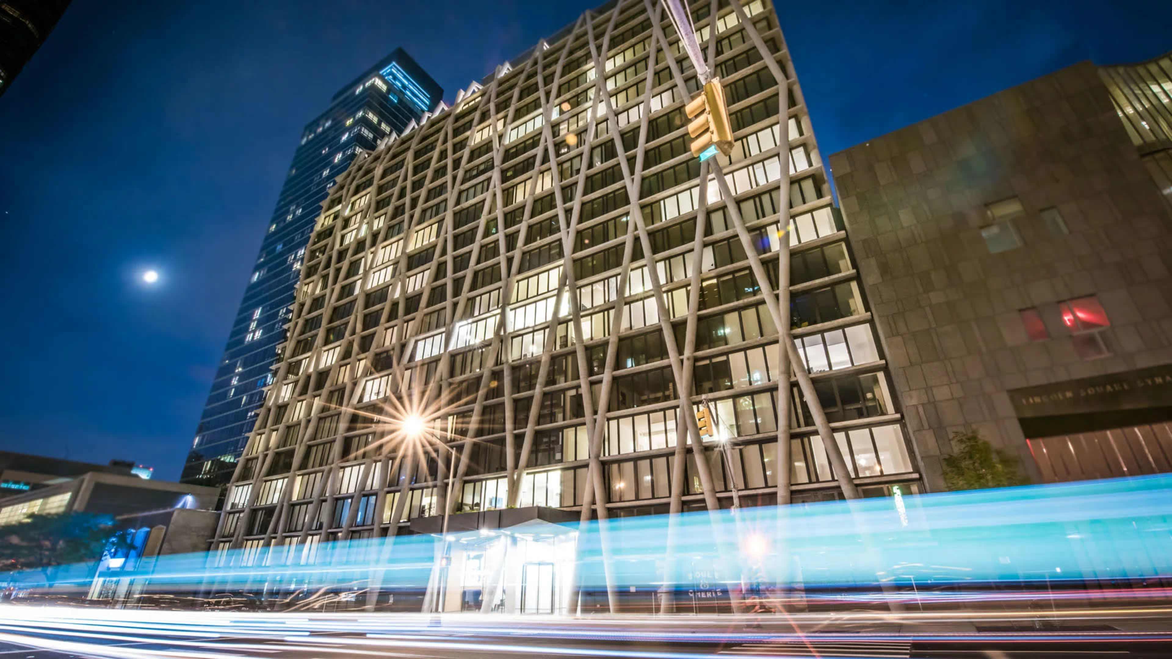 The image shows a modern multi-story building with a geometric facade, illuminated at night, alongside a road with streaks of light from passing vehicles.