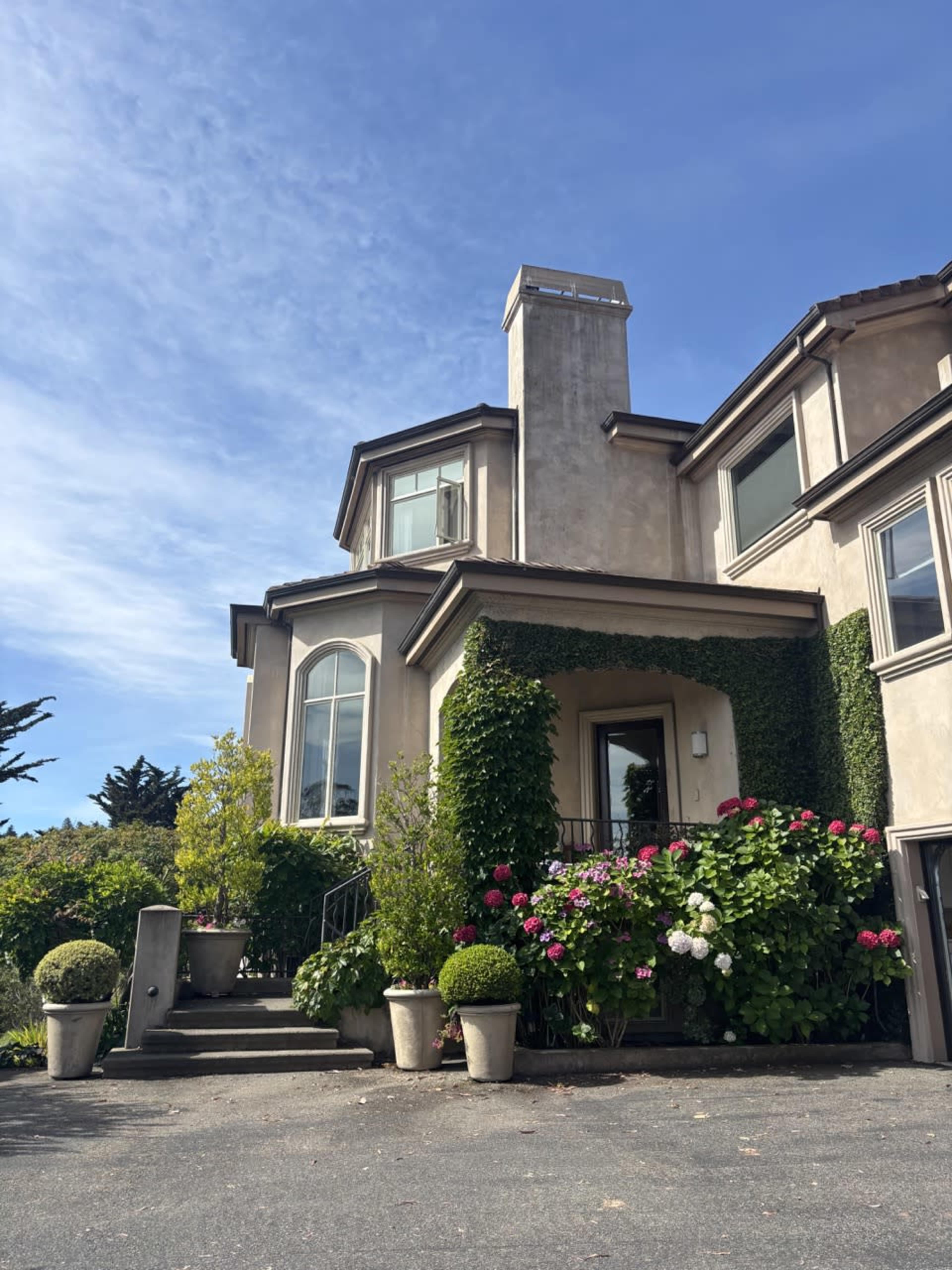 The image shows a two-story house with a stone facade, a prominent chimney, and lush greenery surrounding the entrance, including potted plants and flowering shrubs.
