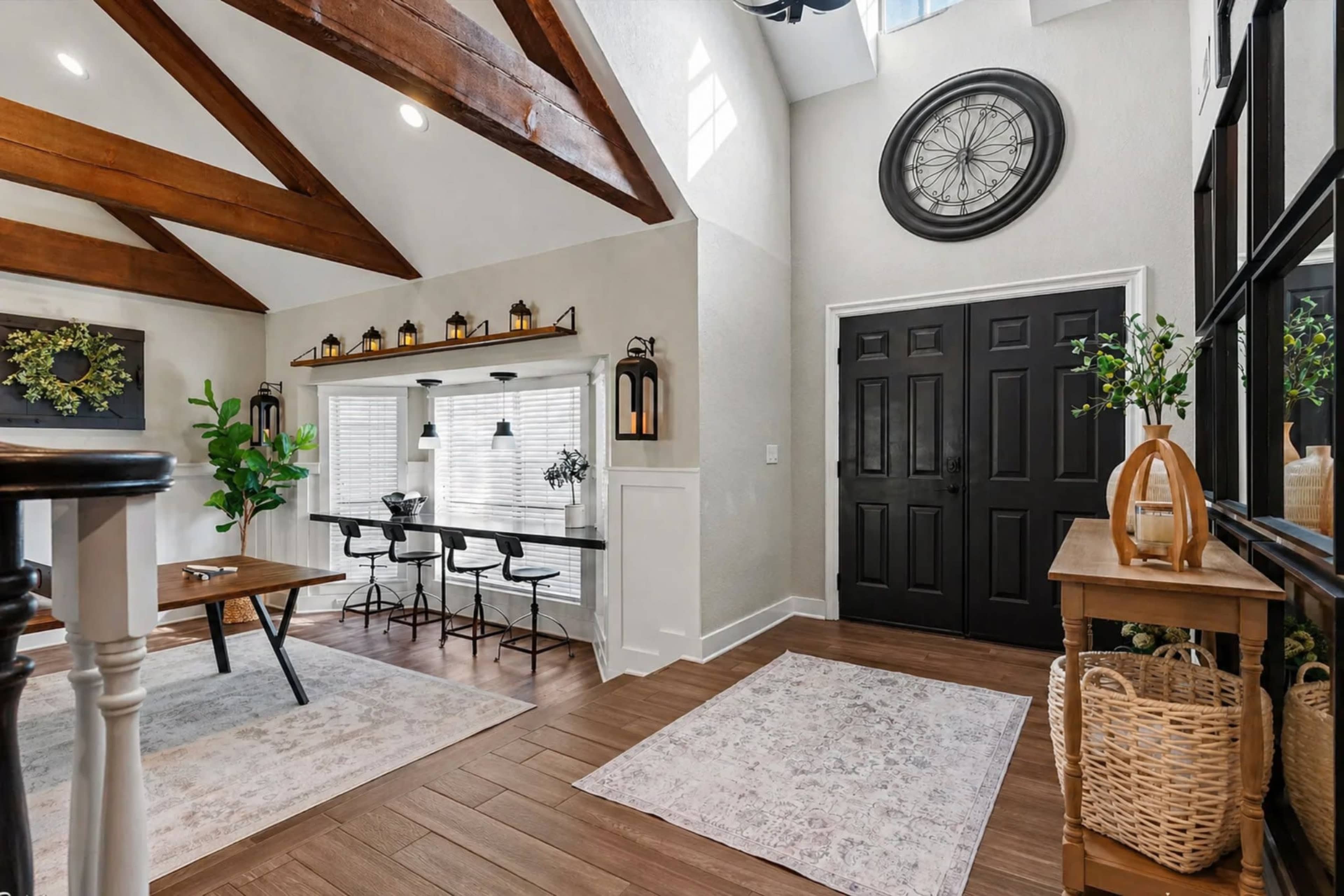 The image shows a spacious entryway featuring a high ceiling with wooden beams, a dark front door, a decorative wall clock, and a dining table with bar stools in a well-lit area.