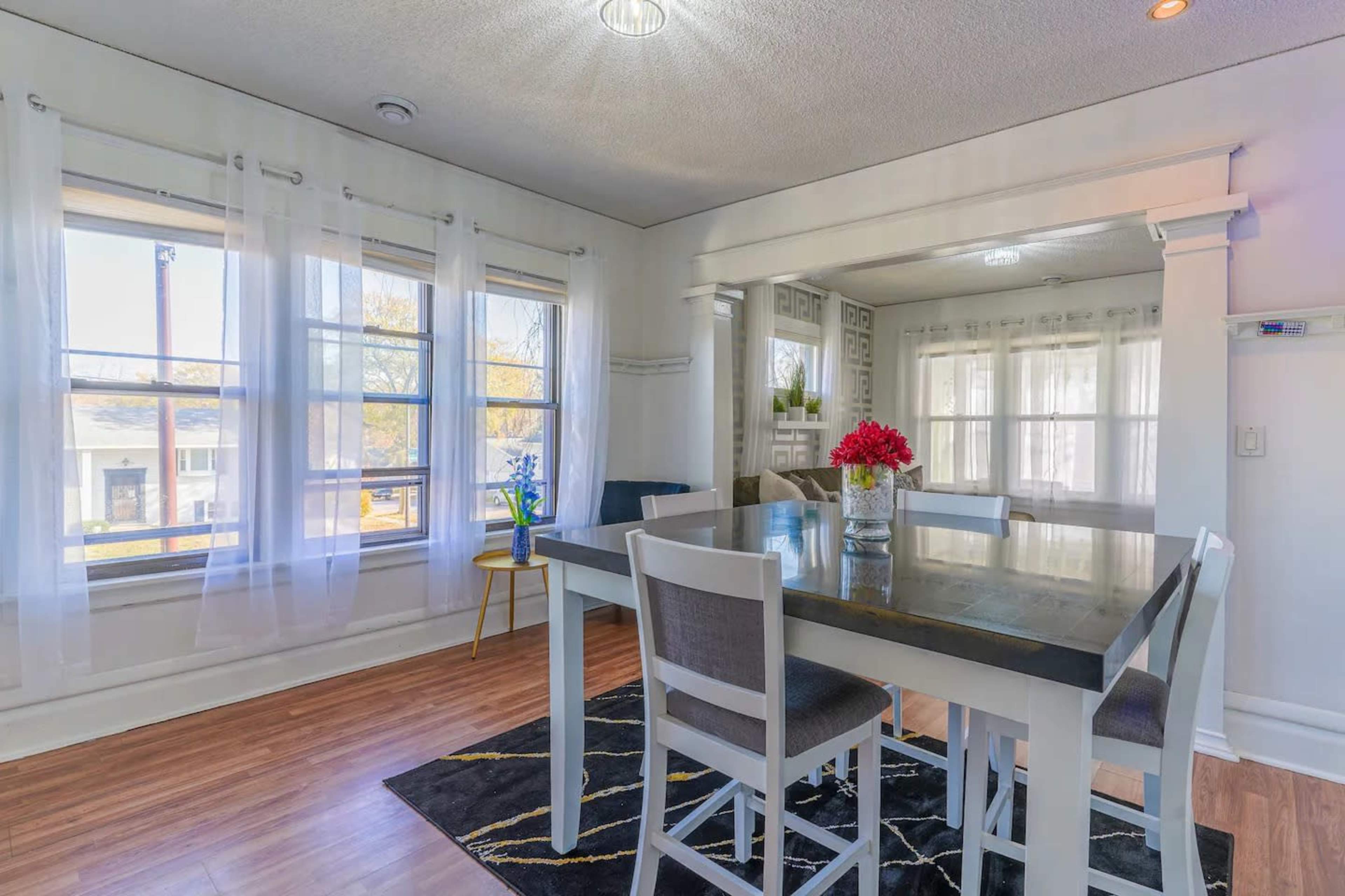 A dining area features a square table surrounded by chairs, with large windows allowing natural light to fill the space.