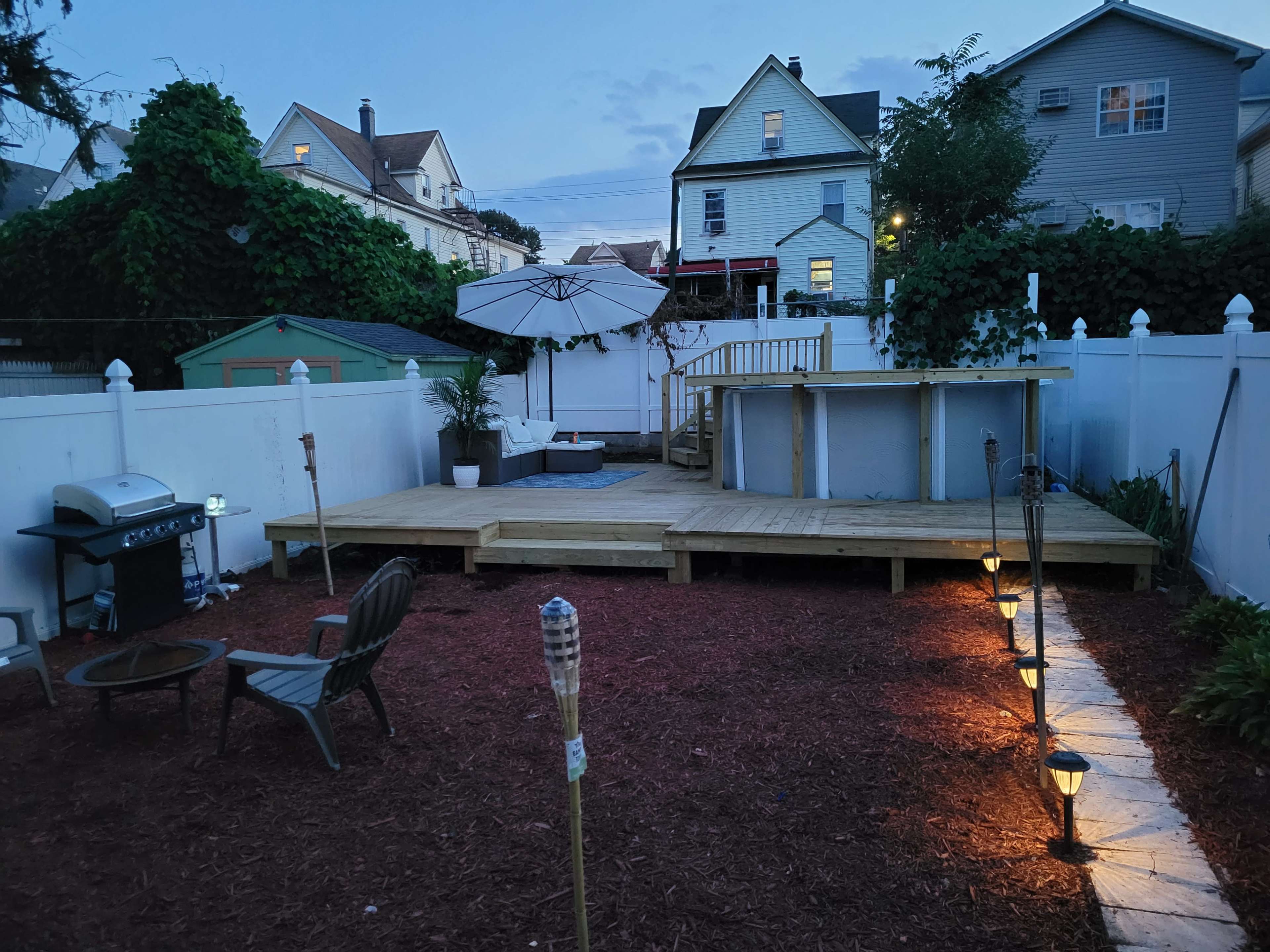 A backyard with a wooden deck, a circular above-ground pool, a grill, patio lights, and a few seating areas, enclosed by white fencing and surrounded by greenery.