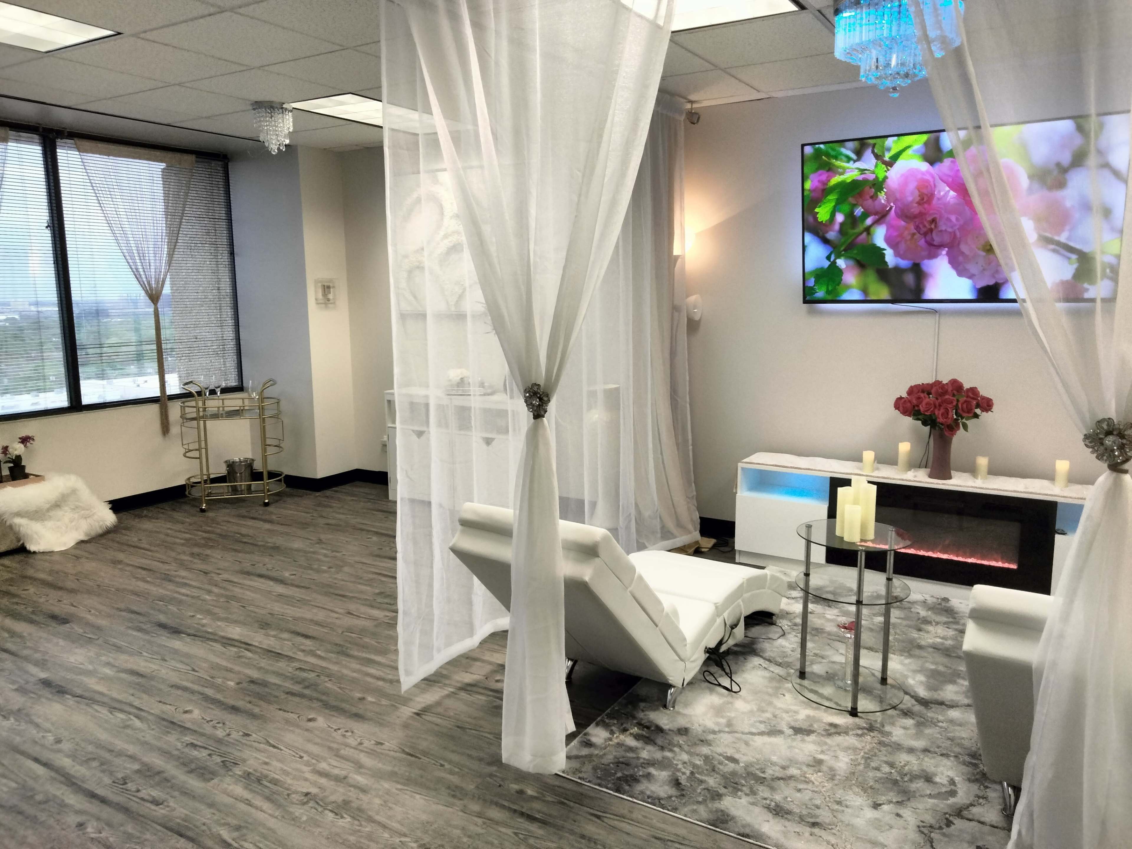 A modern, elegantly designed spa room with a white reclining chair, a circular glass table, and soft lighting, complemented by sheer curtains and a wall-mounted TV displaying floral imagery.