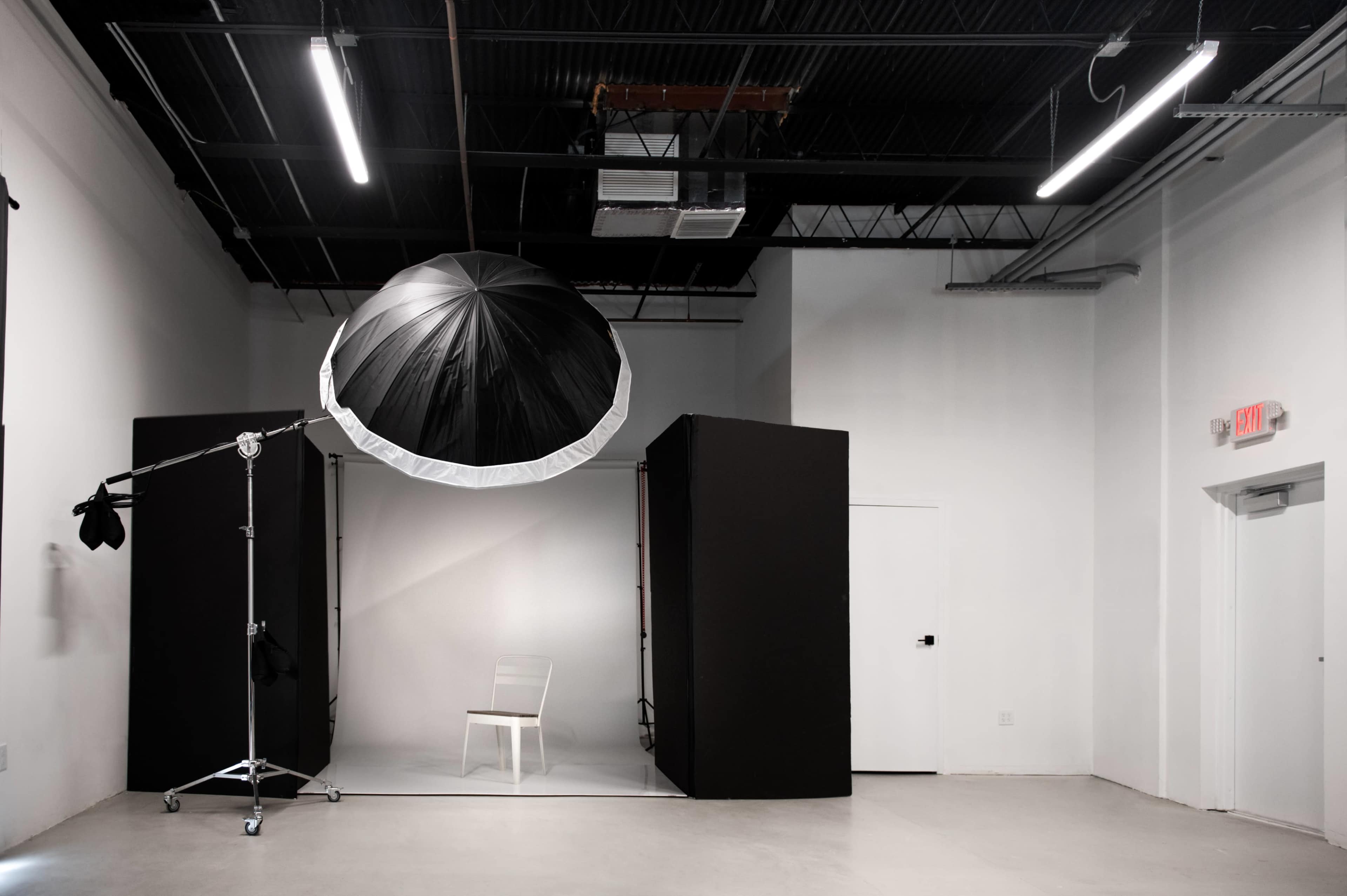 The image shows a photography studio with a large octagonal umbrella light positioned above a white chair in a minimalist setting with dark backdrops.