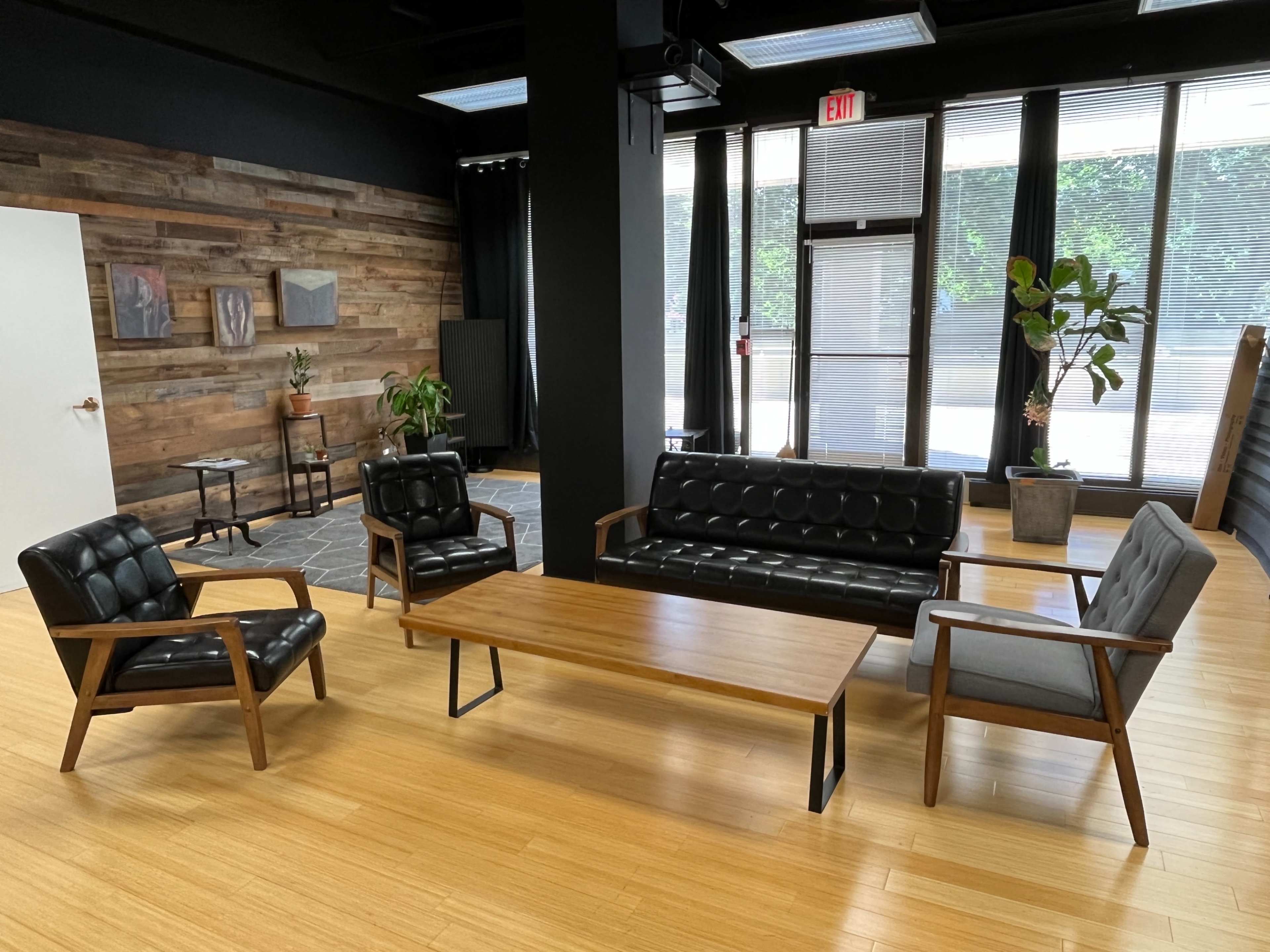 The image shows a modern seating area with a black leather sofa, two upholstered armchairs, a wooden coffee table, and a large indoor plant, all set against a backdrop of wooden paneling and large windows.