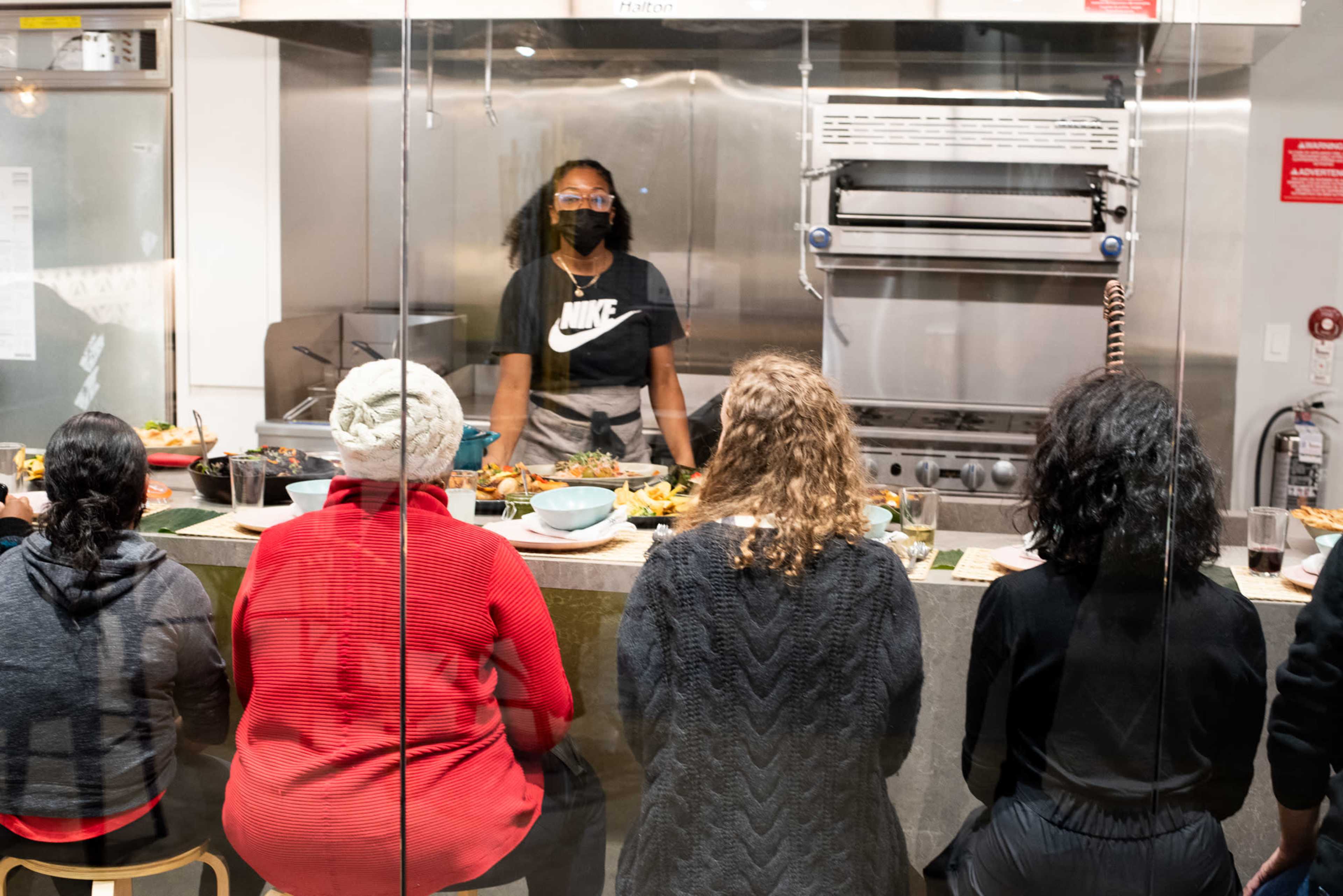 A chef stands behind a kitchen counter preparing dishes while a group of diners observes from the other side of a glass partition.