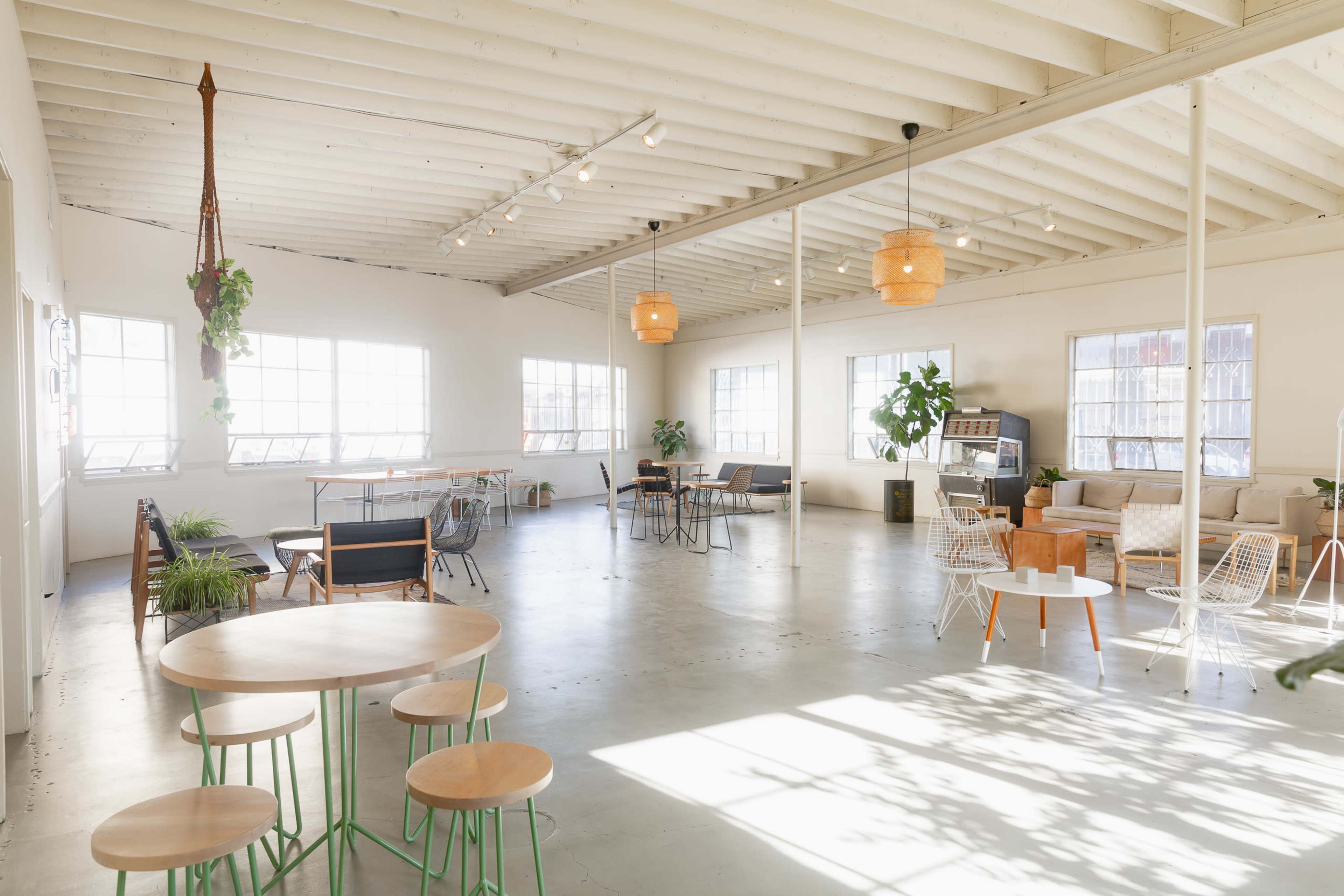 The image shows a spacious, well-lit interior with wooden beams, various tables and chairs, and large windows allowing natural light to fill the space.