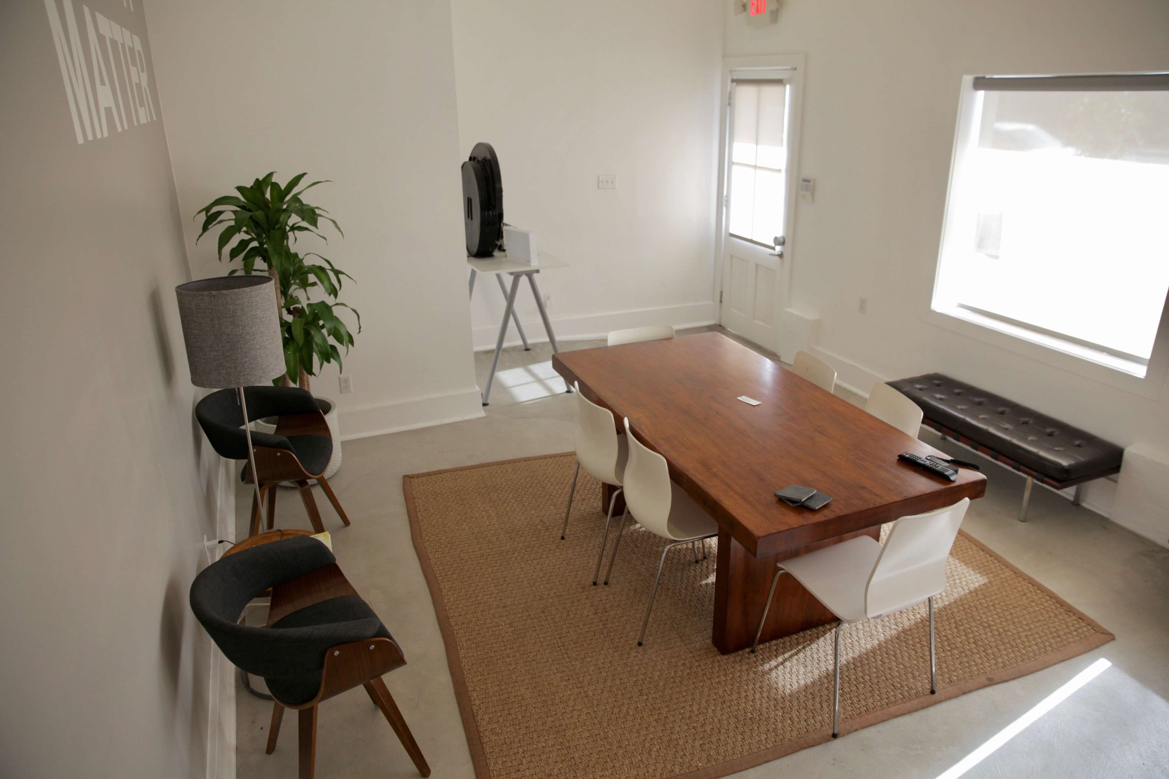 A modern conference room features a wooden table surrounded by white chairs, a large plant in the corner, and a seating area with a couch and lamp.