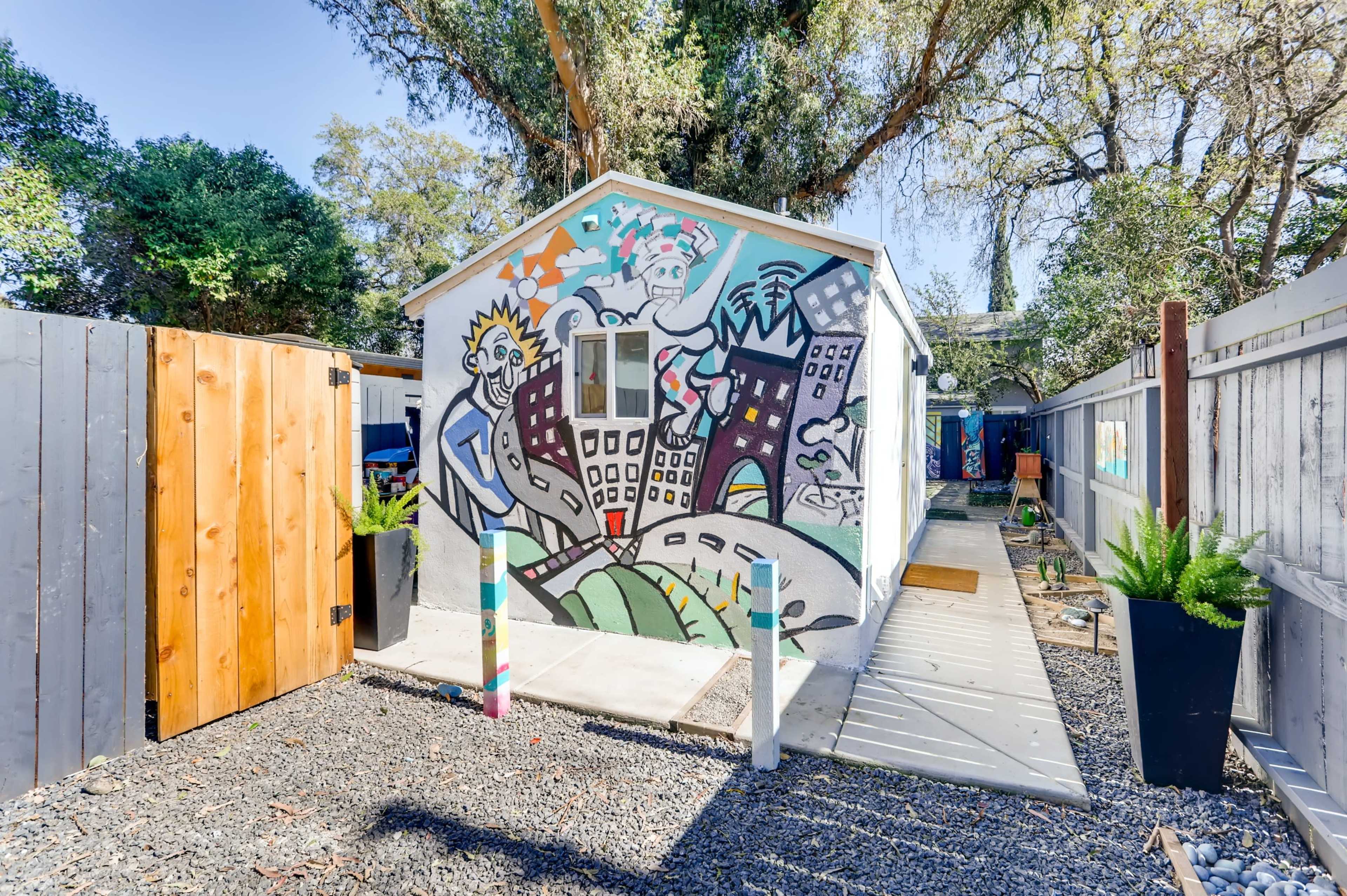 The scene depicts a colorful mural on a small building surrounded by a gravel pathway and landscaping elements in a garden setting.
