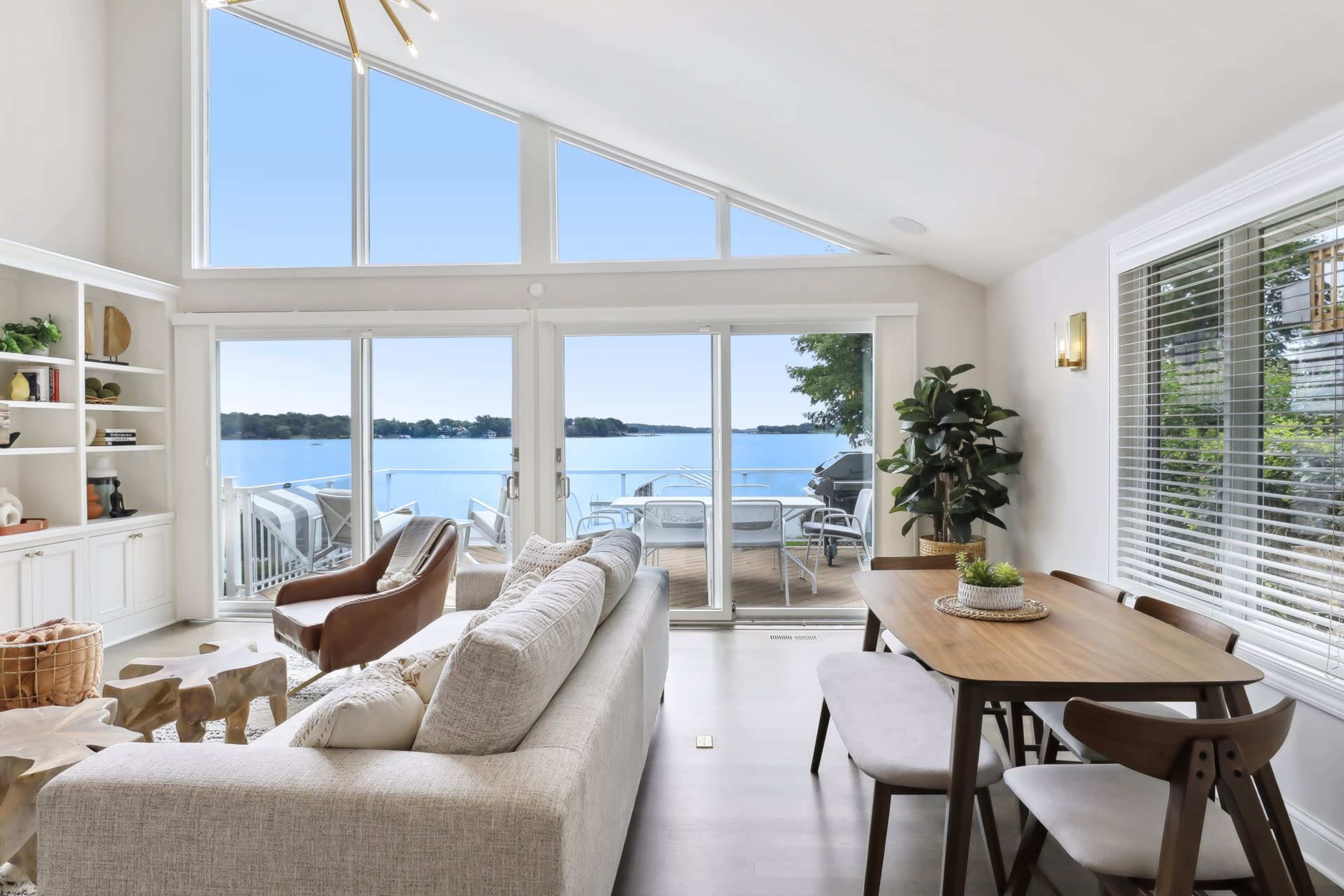 A bright, modern living area features a large window overlooking a lake, with comfortable seating and a dining table nearby.