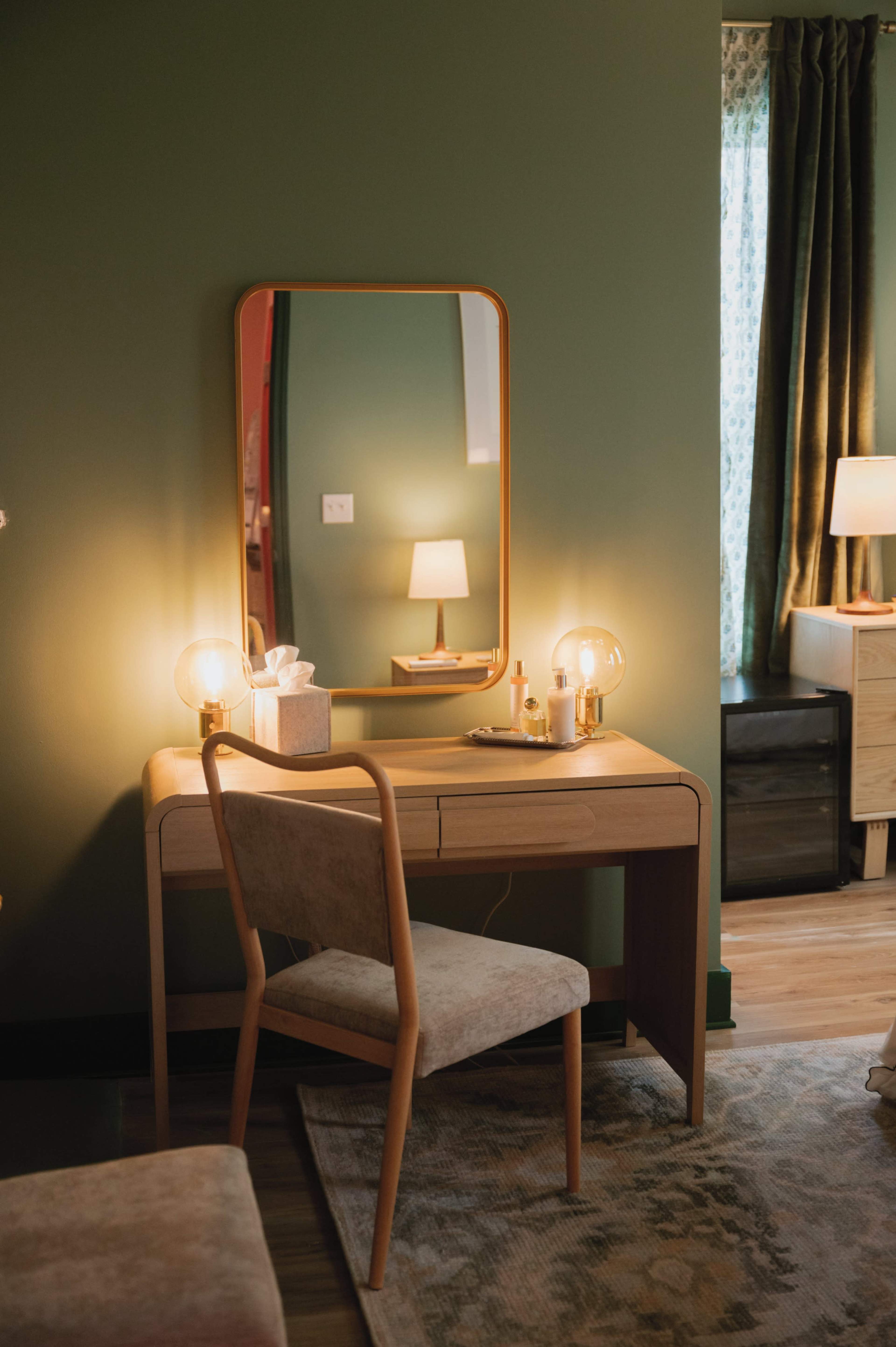 A simple wooden vanity with a large mirror and soft lighting is positioned against a green wall in a well-decorated bedroom.