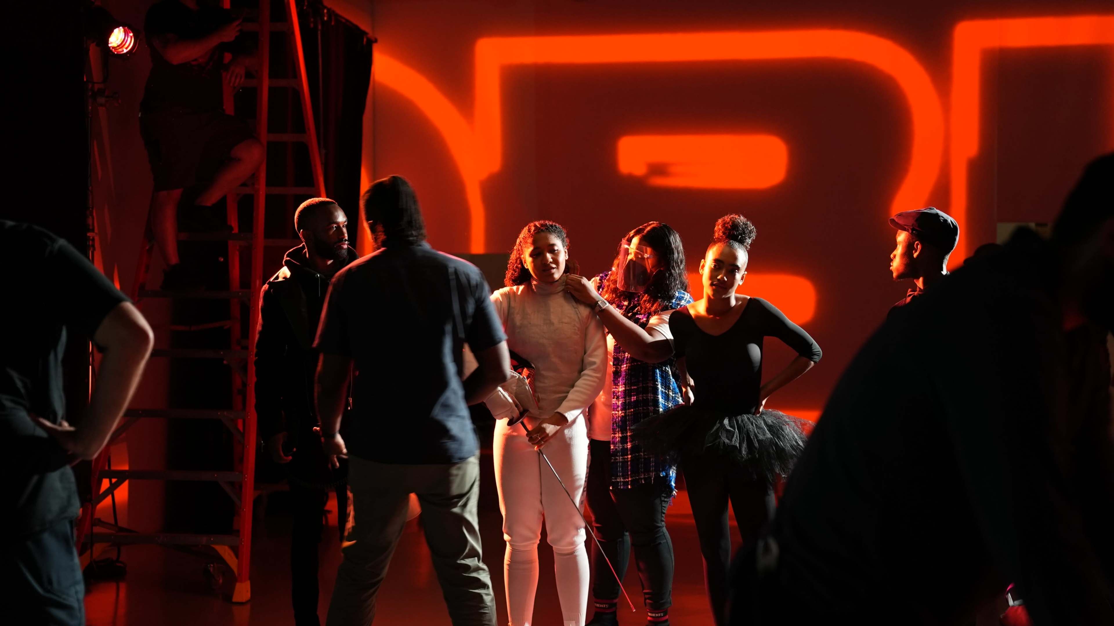 A group of people stands on a stage with a large illuminated backdrop while others work on equipment in the background.