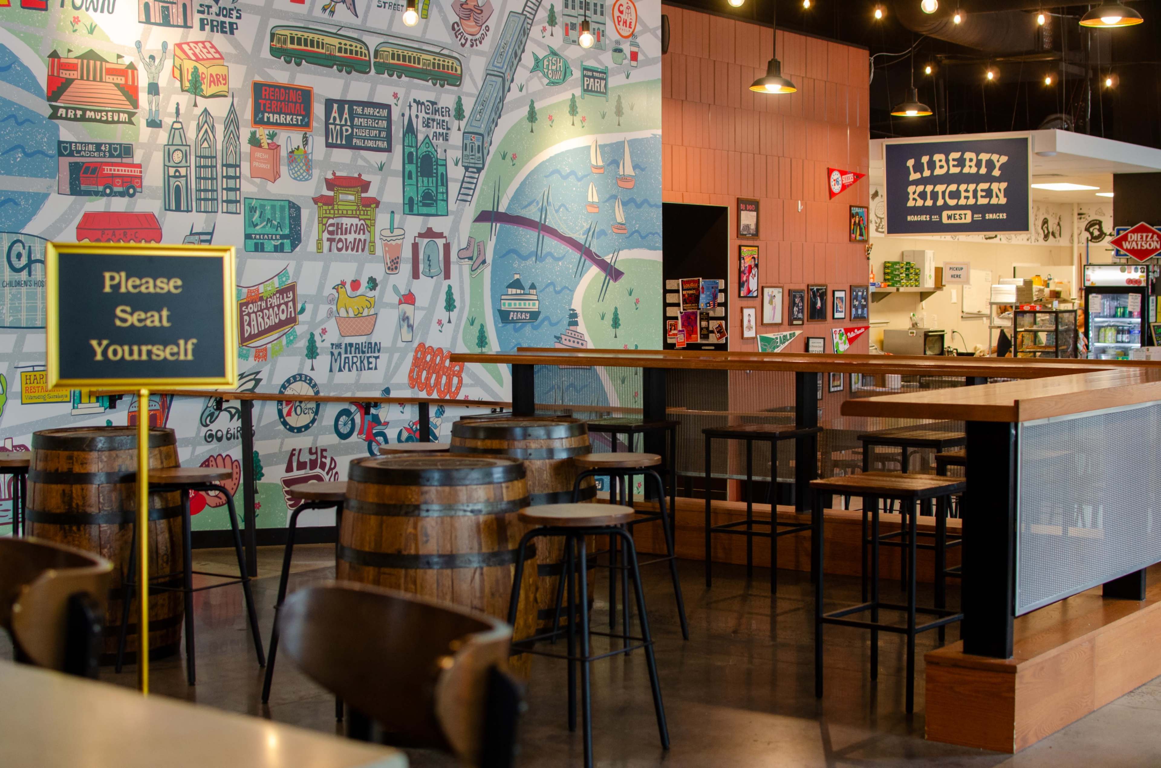 The image shows a brightly decorated interior of a restaurant with wooden tables, tall stools, and a mural featuring various city landmarks on the wall.