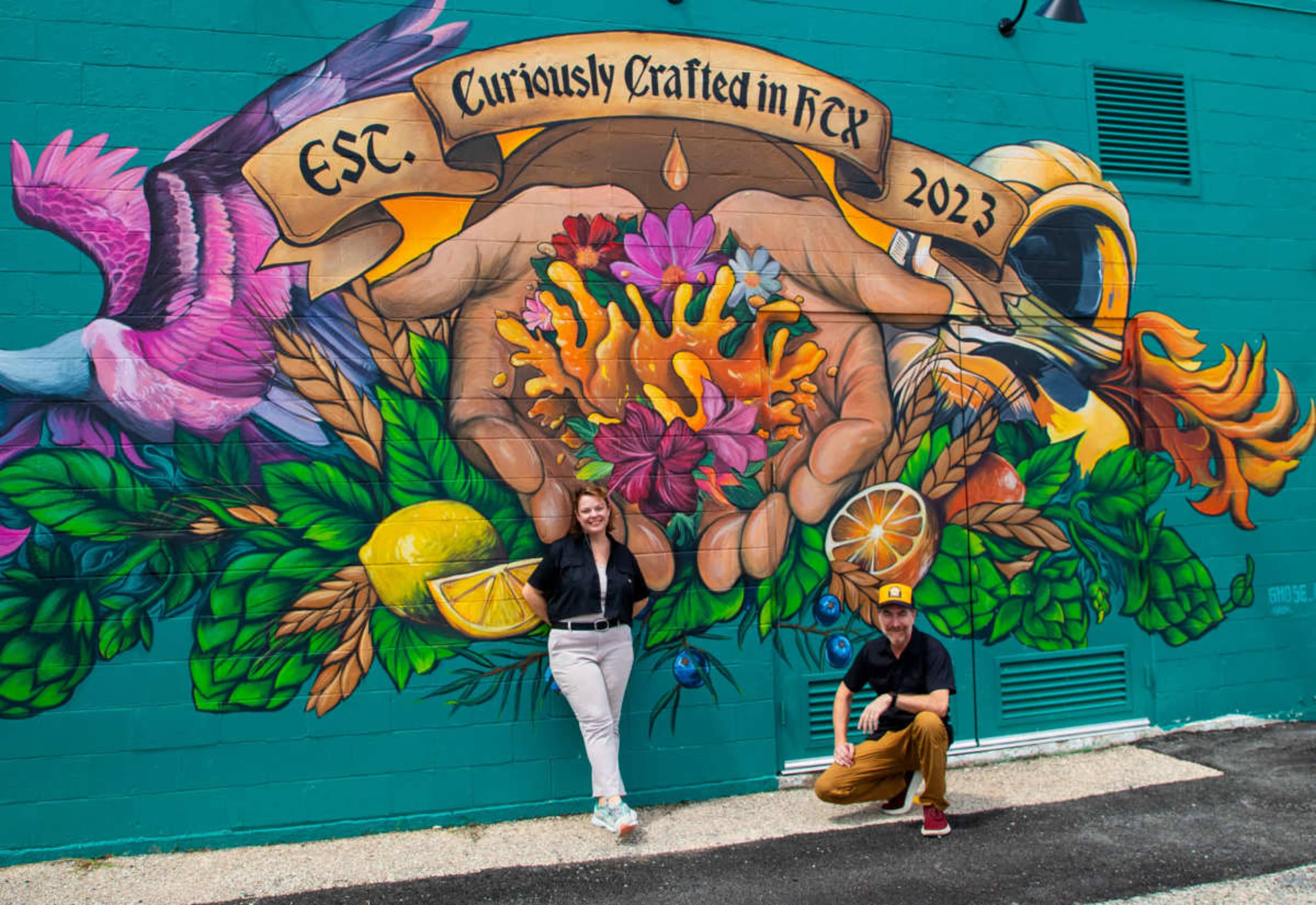 A man and a woman pose in front of a colorful mural featuring hands holding vibrant flowers and fruits, with the text "EST. Curiously Crafted in HTX 2023" above.