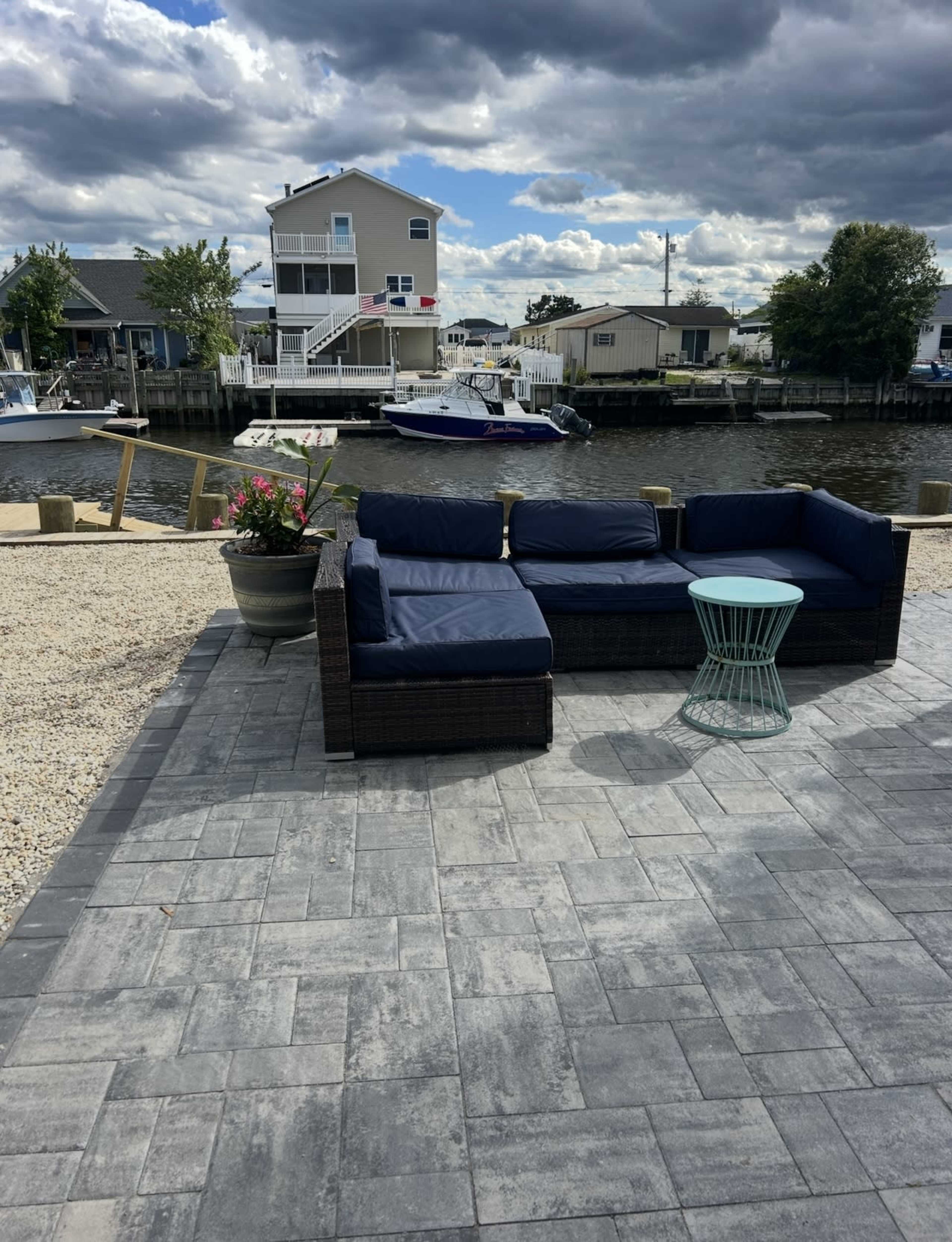 A patio area features a navy blue sectional sofa and a light teal coffee table overlooking a canal with boats and houses in the background.