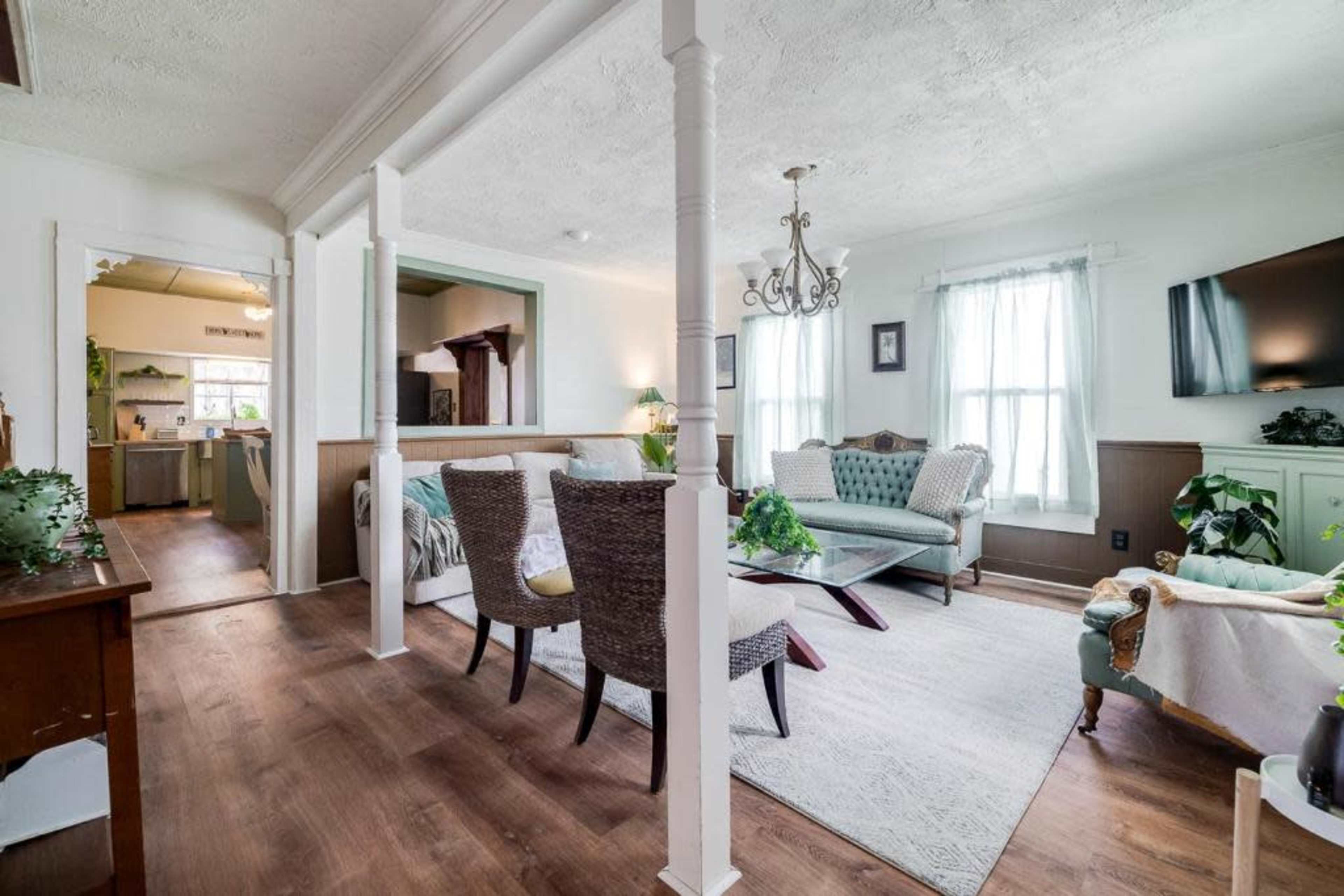 A cozy living room with a mix of upholstered seating, white walls, large windows, and a doorway leading to a kitchen area.