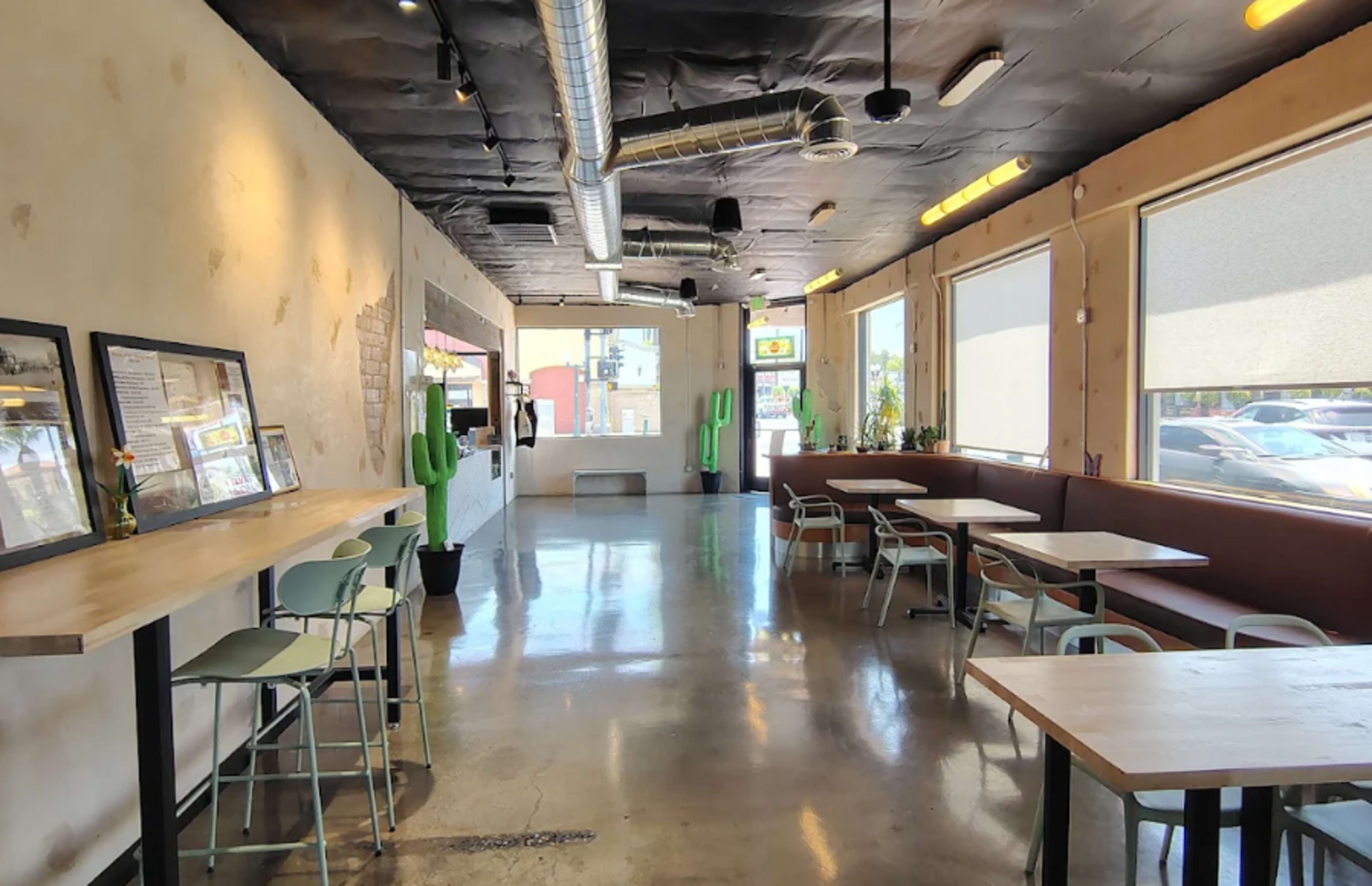 The image shows the interior of a modern restaurant with a sleek design, featuring sparse seating, large windows, and green cactus decorations.