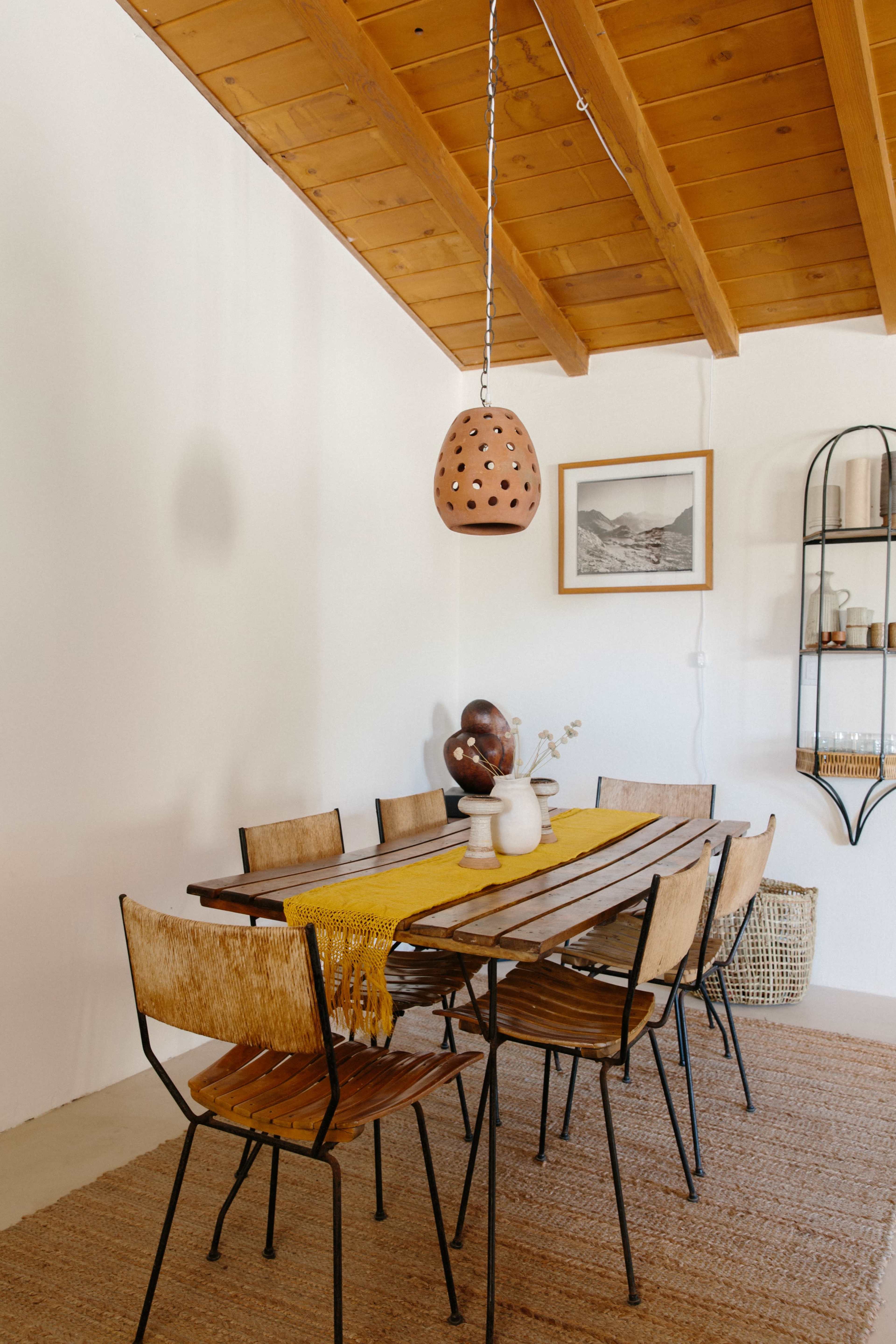 A wooden dining table with metal chairs is set beneath a wooden ceiling, accompanied by a decorative pendant light and shelves displaying various items.